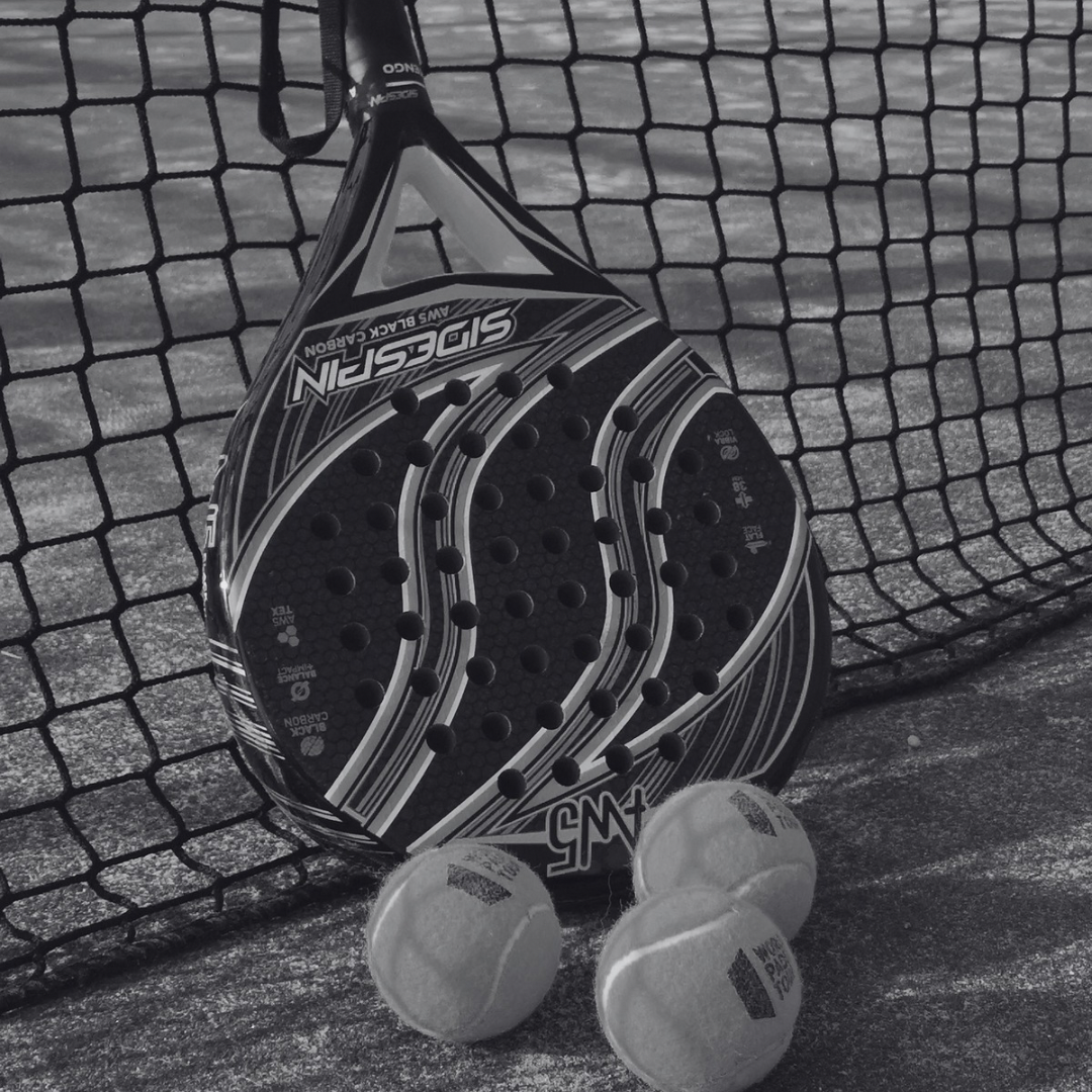 A padel racket leaning against a net with three tennis balls lying in front of it on a clay court.