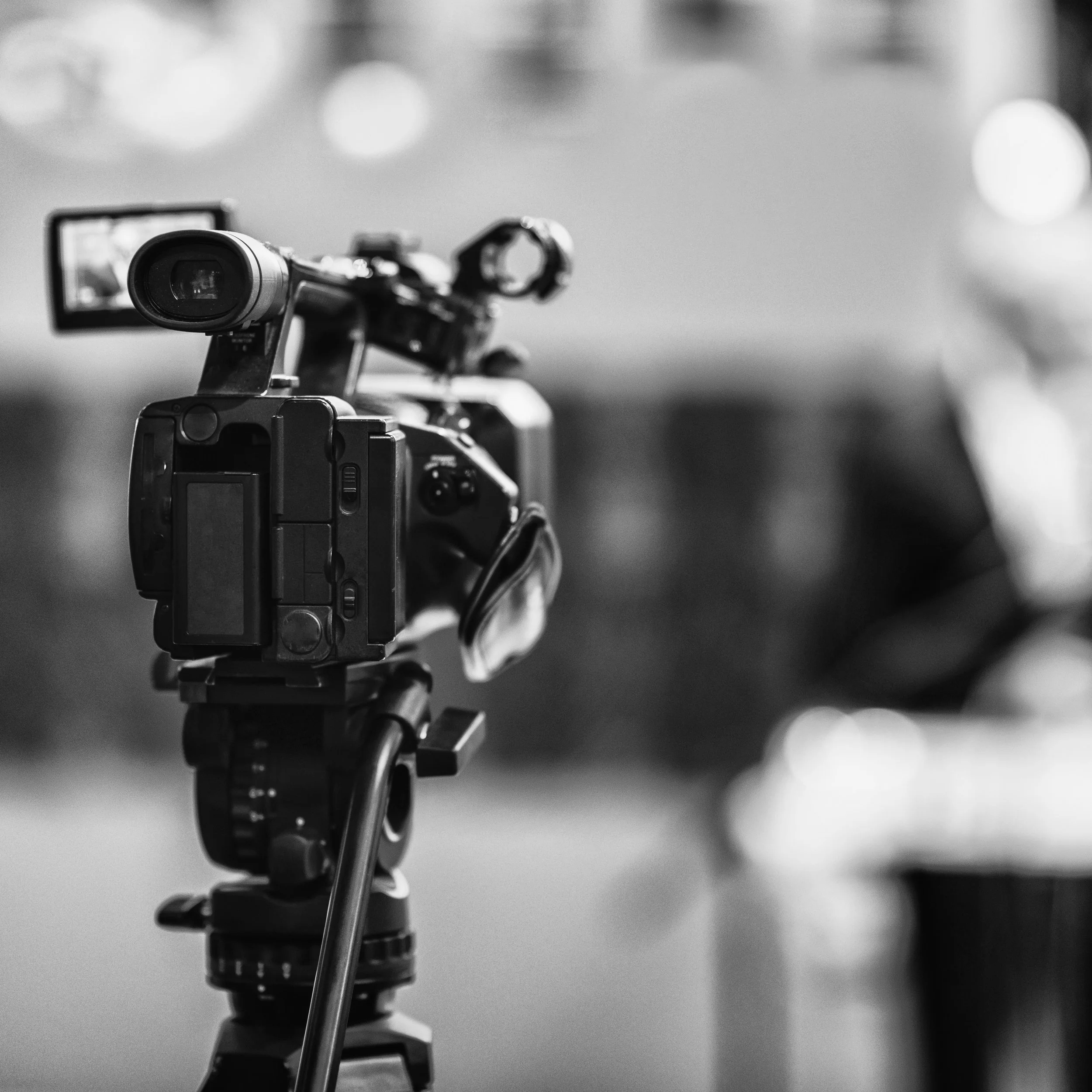 A black-and-white photograph of a professional video camera on a tripod, aimed forward. The background is blurred with circular light bokeh effects.