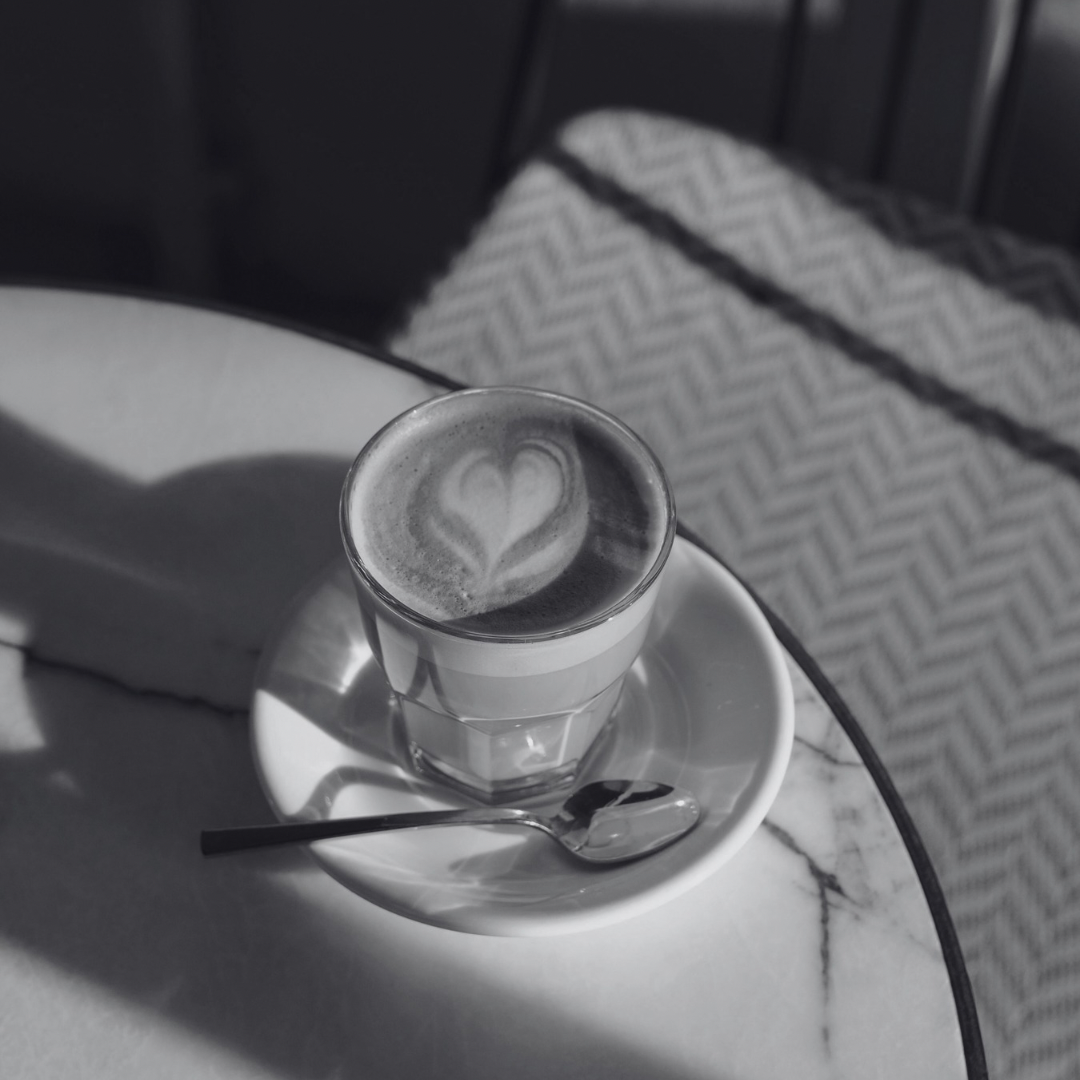 A glass of coffee with latte art in the shape of a heart on top, served on a saucer with a teaspoon, on a white table.
