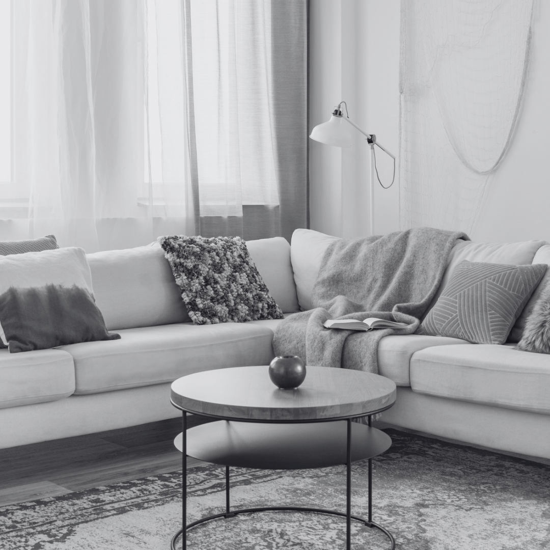 Black and white photo of a modern living room with a white sofa decorated with various textured pillows and throws. A round coffee table with a small dark vase in the center is in front of the sofa. Sheer curtains cover the windows, and a wall-mounted lamp is above the sofa.