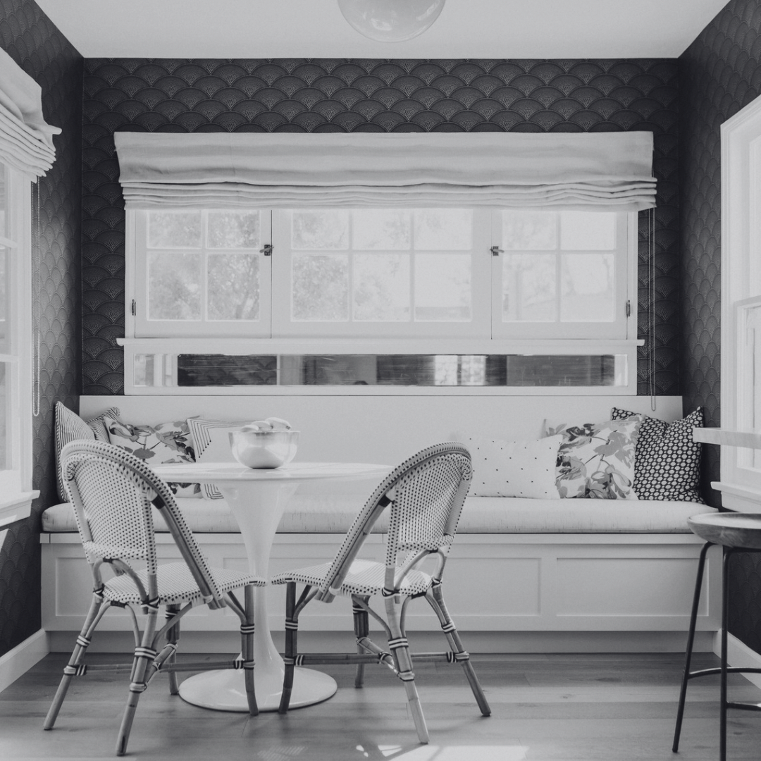 A black and white photo of a cozy dining nook with a built-in bench under a large window, which has decorative pillows and a white seat cushion. There is a small round table with a bowl on top and three cane-backed chairs around it. The walls have a patterned wallpaper, and the room is well-lit with natural light from the windows.