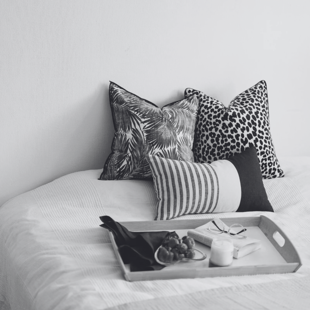 A bed with four decorative pillows and a tray with a candle, sunglasses, and fruit.