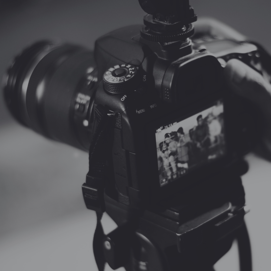 A black and white photograph of a digital camera on a tripod, focused on the camera's LCD screen which displays a photo of several people.