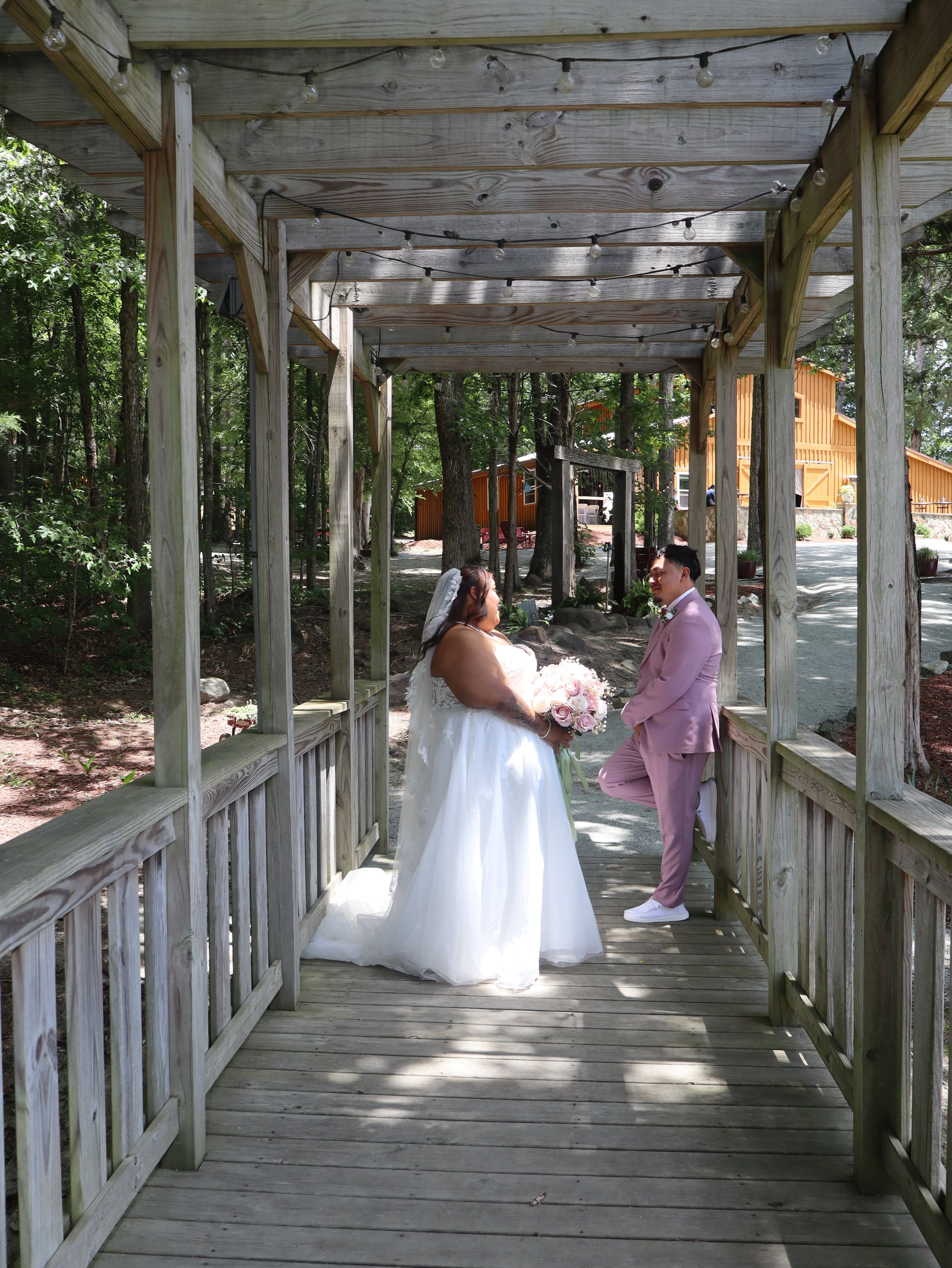 Bride and groom standing under a wooden gazebo during their outdoor wedding. The bride is in a white gown holding a bouquet, and the groom is in a light pink suit. Green trees and wooden buildings are visible in the background.