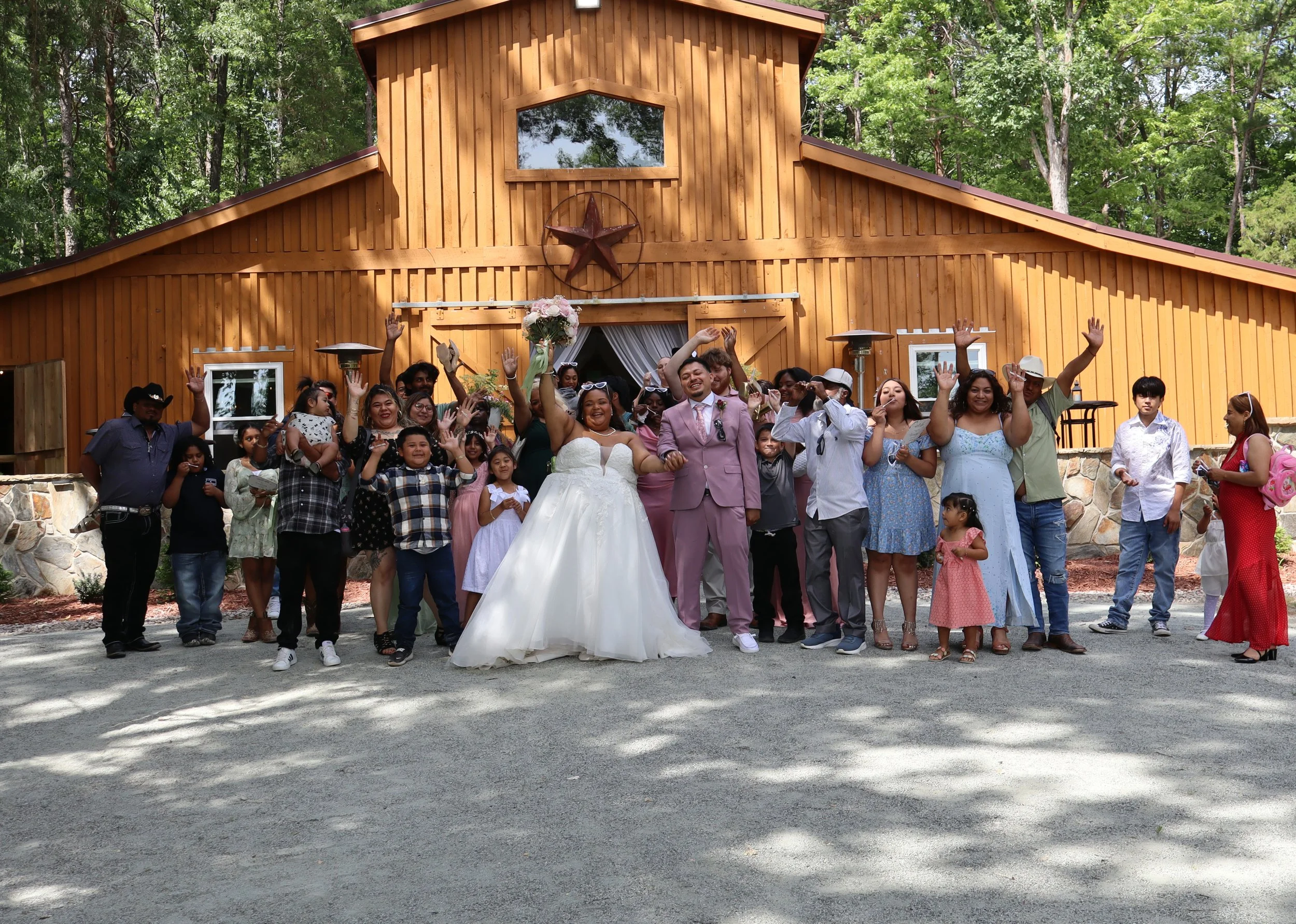 Group of people celebrating in front of a wooden barn, some waving and smiling, a bride in a wedding dress and groom in a pink suit in the center, surrounded by family and friends.