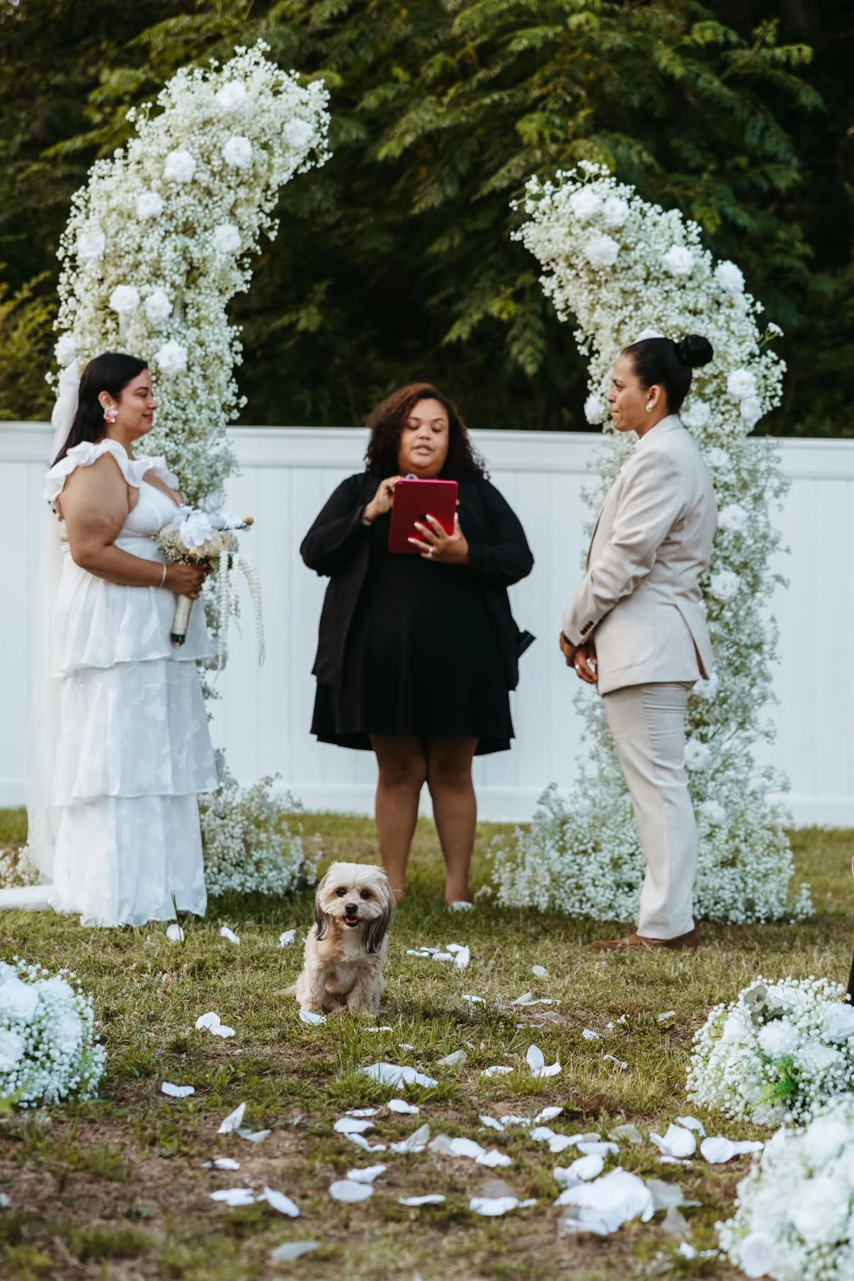 A wedding ceremony outdoors with two women, one in a white dress holding a bouquet, and another in a beige suit, standing under a floral arch. An officiant is reading from a document, and a small dog sits on the grass among scattered flower petals, with trees in the background.