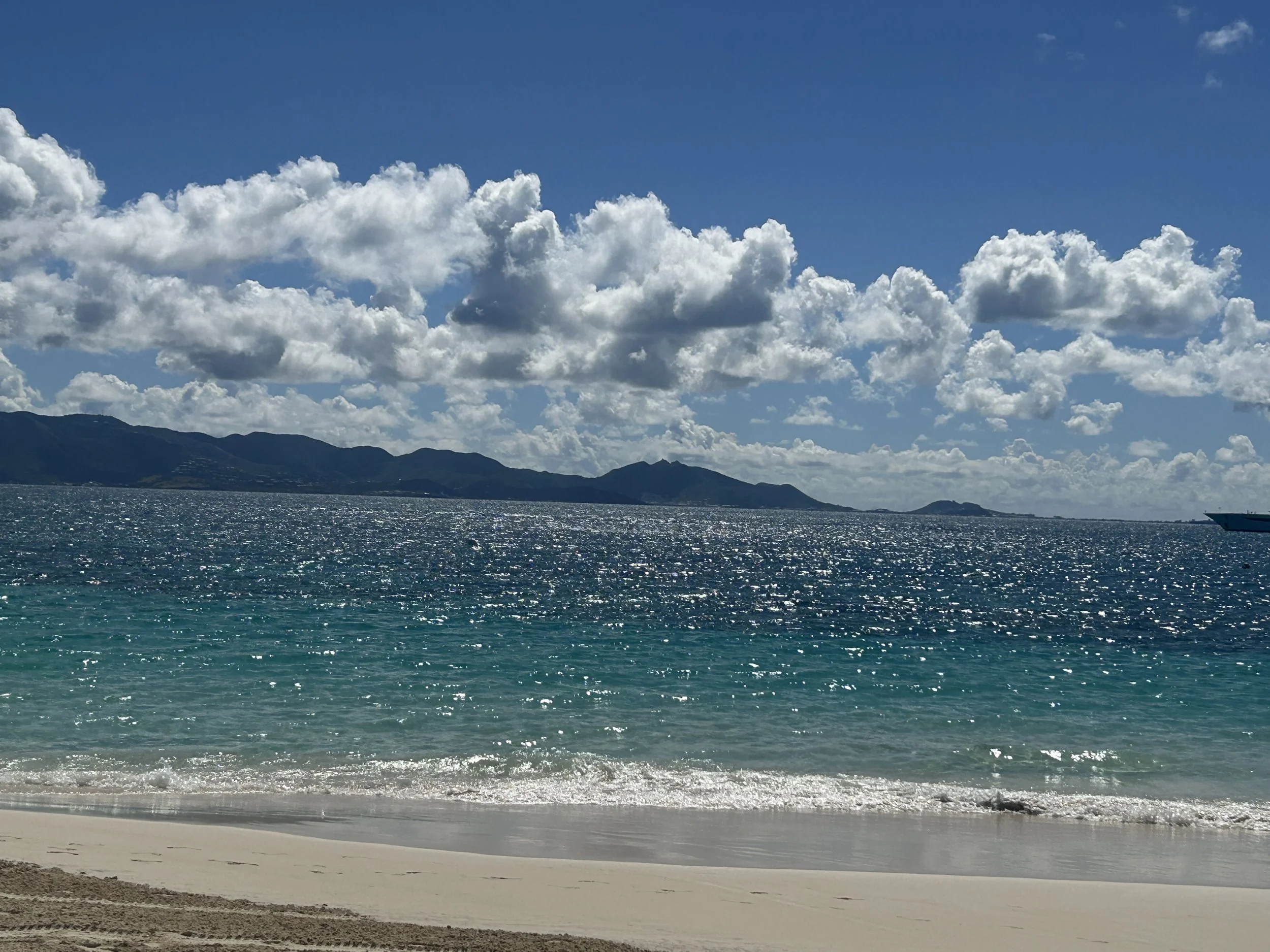 A beach scene with clear blue water, a mountain range in the distance, partly cloudy sky, and sandy shoreline in the foreground.