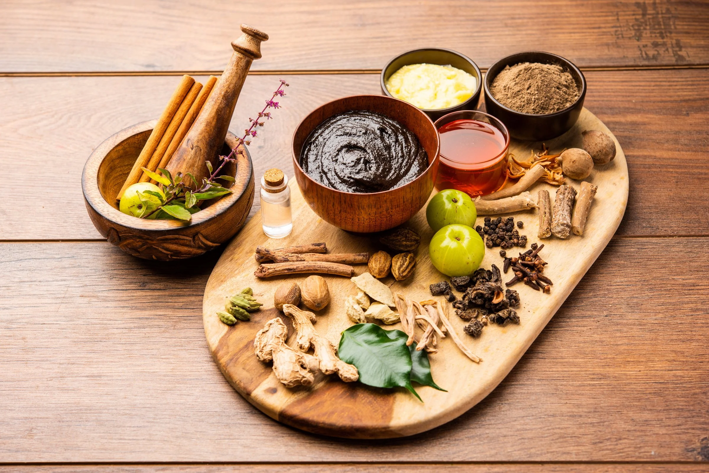 A wooden board with various spices, herbs, and natural ingredients, including cinnamon sticks, cardamom, dried herbs, and small bowls containing creams, powders, and liquids, arranged for culinary or medicinal purposes.