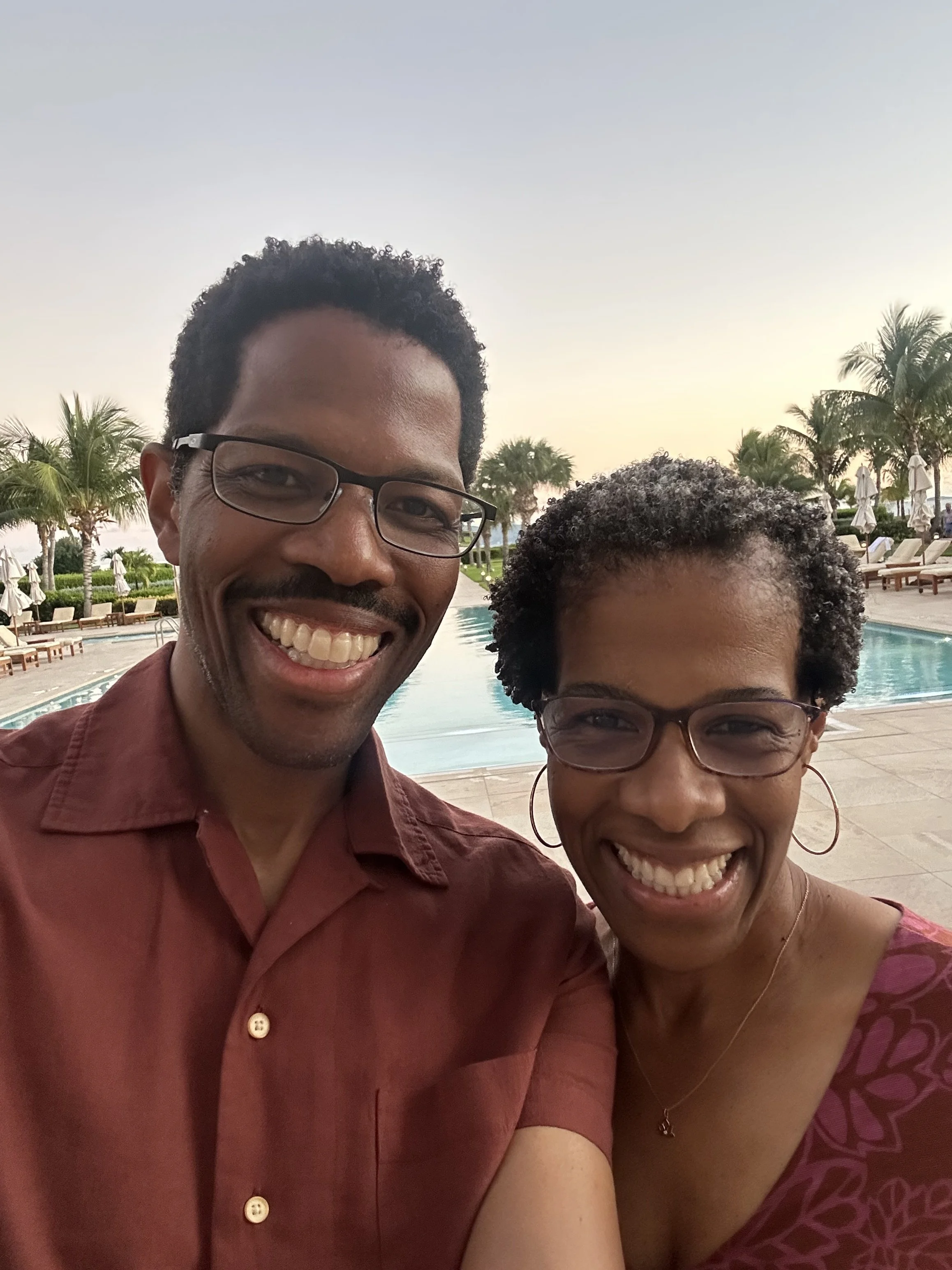 Smiling man and woman taking a selfie by a poolside with palm trees and lounge chairs in background during sunset.