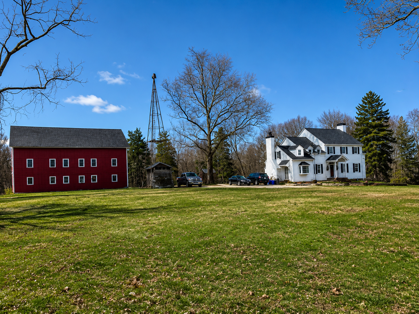 A rural scene features a large white house with black shutters, a red barn, a tall leafless tree, several parked cars, and a weather vane on a small shed, under a bright blue sky with some clouds.