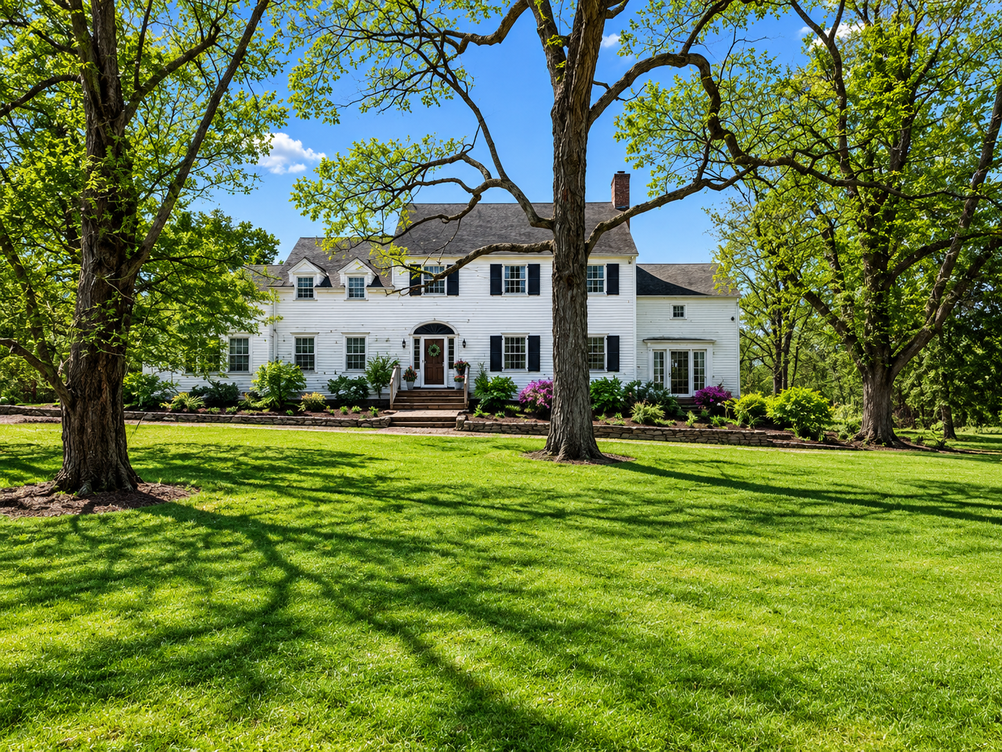 A large white house with black shutters and a front porch, surrounded by green trees and a well-maintained lawn on a sunny day.