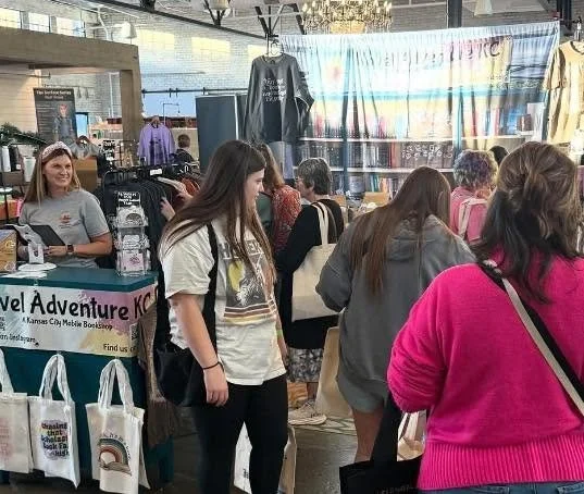 People browsing booths at an indoor event, with a table displaying books and merchandise, and a banner in the background with colorful text.