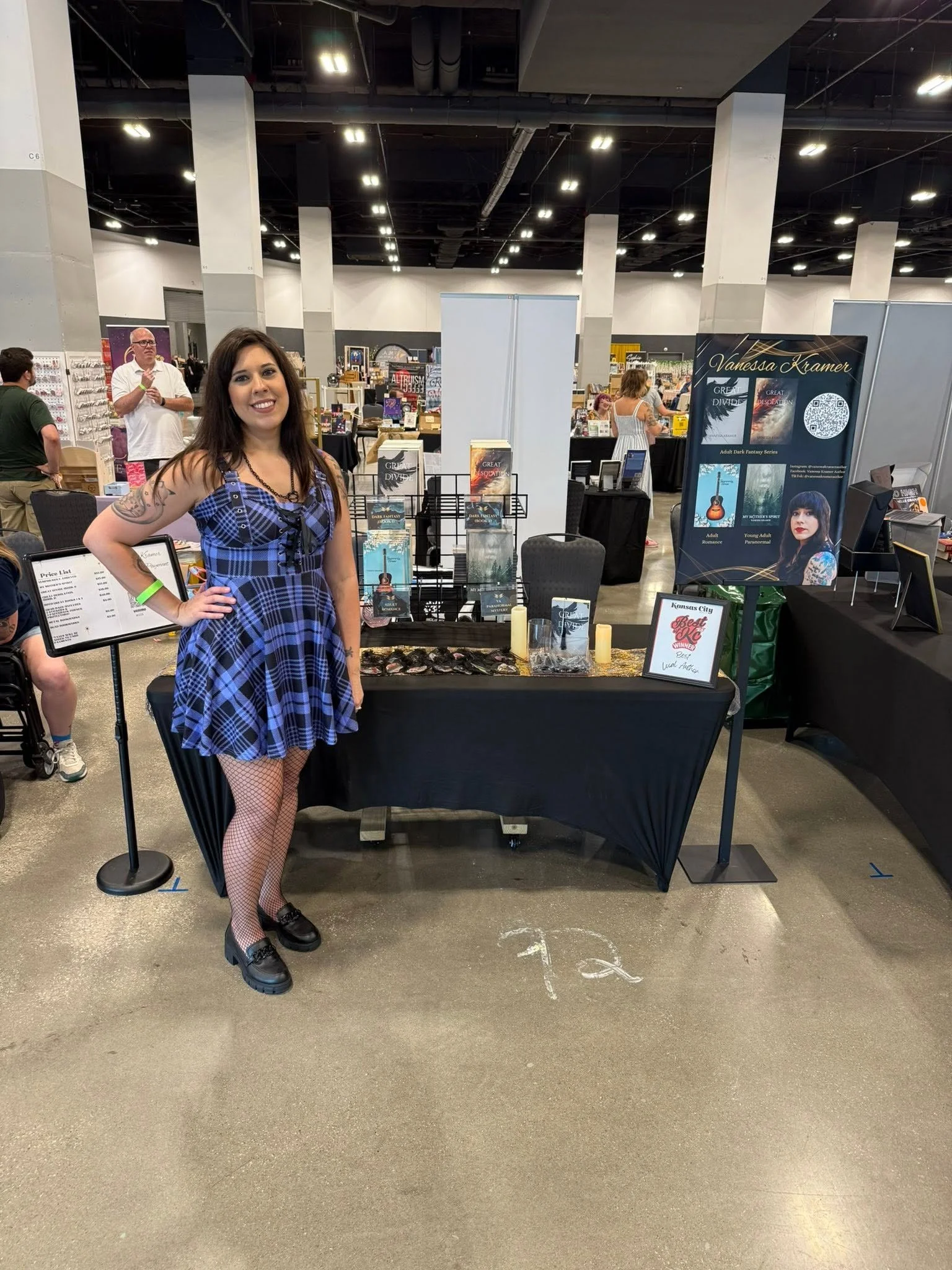 A woman with long dark hair, wearing a blue plaid dress, fishnet stockings, and black shoes, standing in front of a booth at a convention or expo, smiling at the camera.