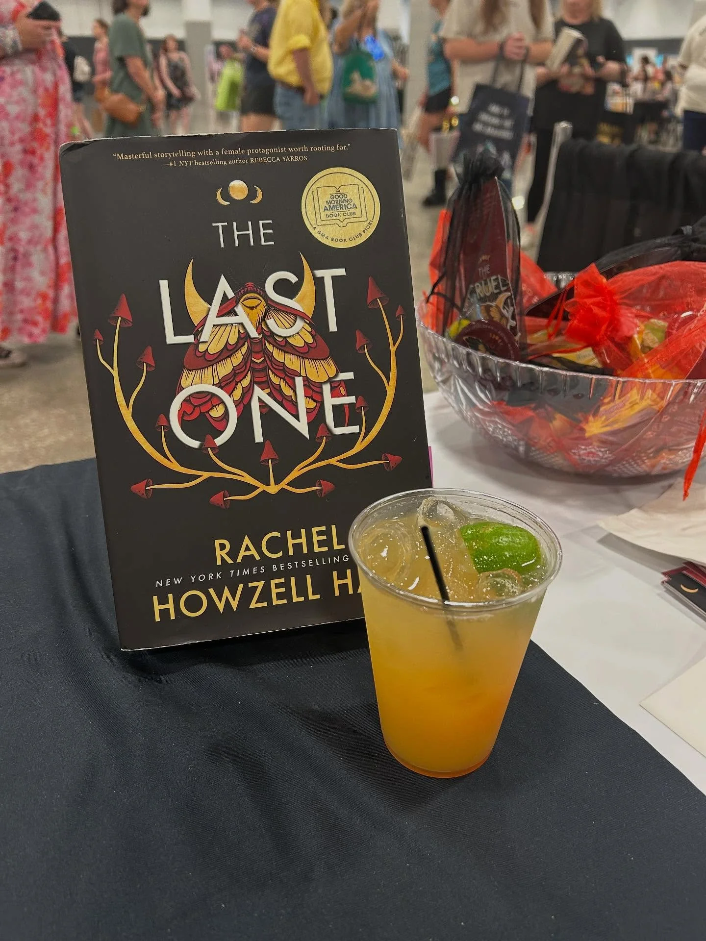 A copy of the book "The Last One" by Rachel Howzell Hall, a glass of yellow beverage with lime and ice, a bowl of candies in orange mesh bags, and people in line in a bookstore or event setting.