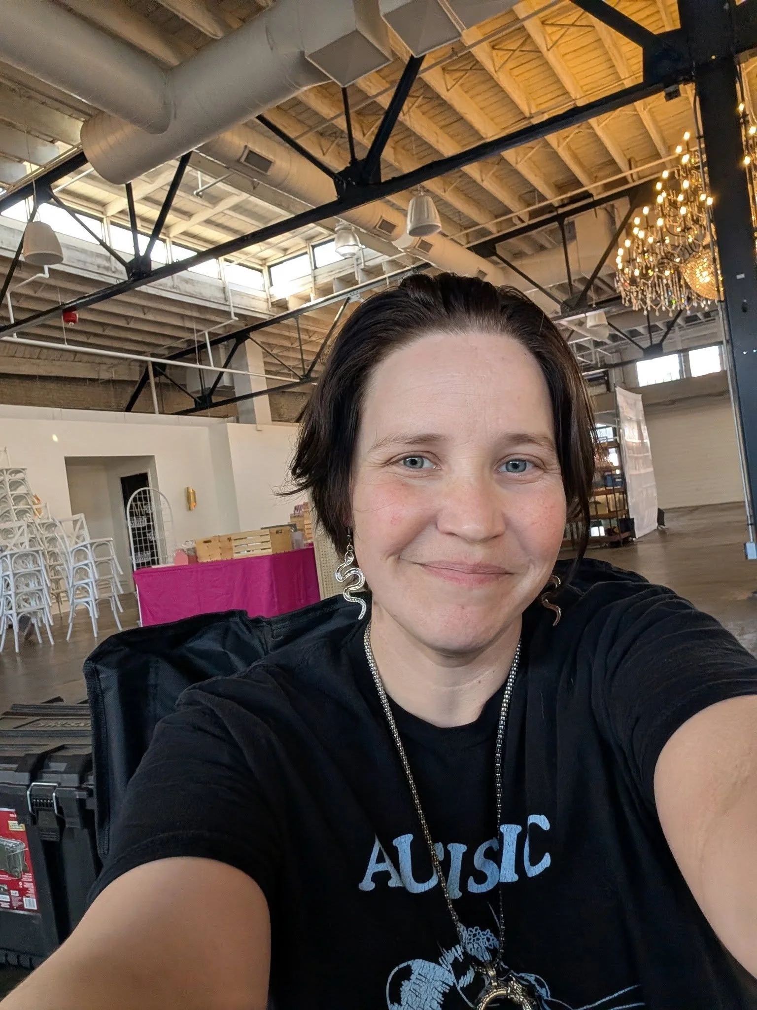 A woman taking a selfie in a spacious indoor setting with high ceilings, exposed beams, and a chandelier, smiling with visible blue eyes and short dark hair, wearing a black T-shirt and jewelry.