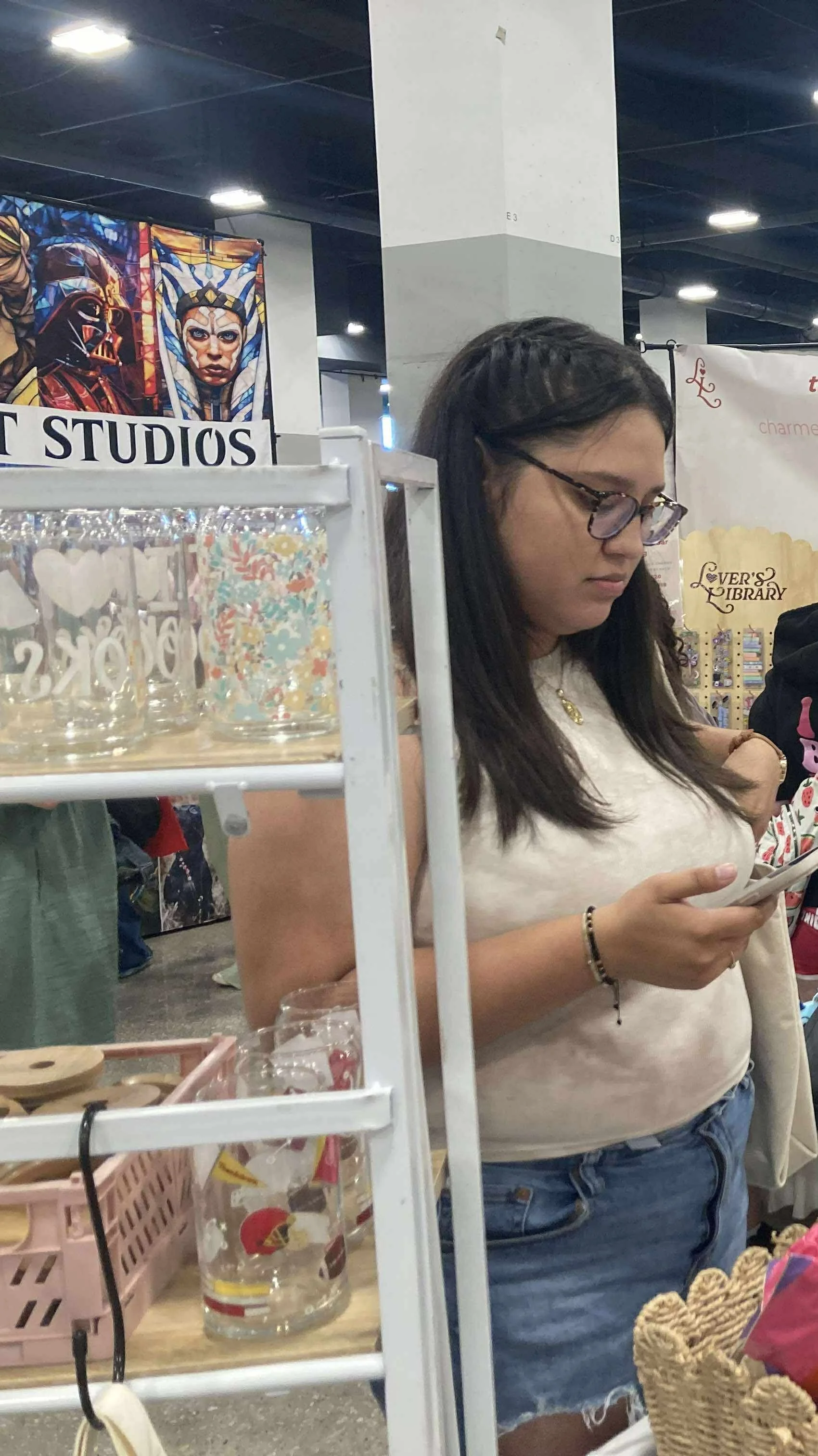 A woman with long dark hair, glasses, wearing a white t-shirt and denim shorts, standing in a store and looking at her phone. Behind her are various booths and displays.