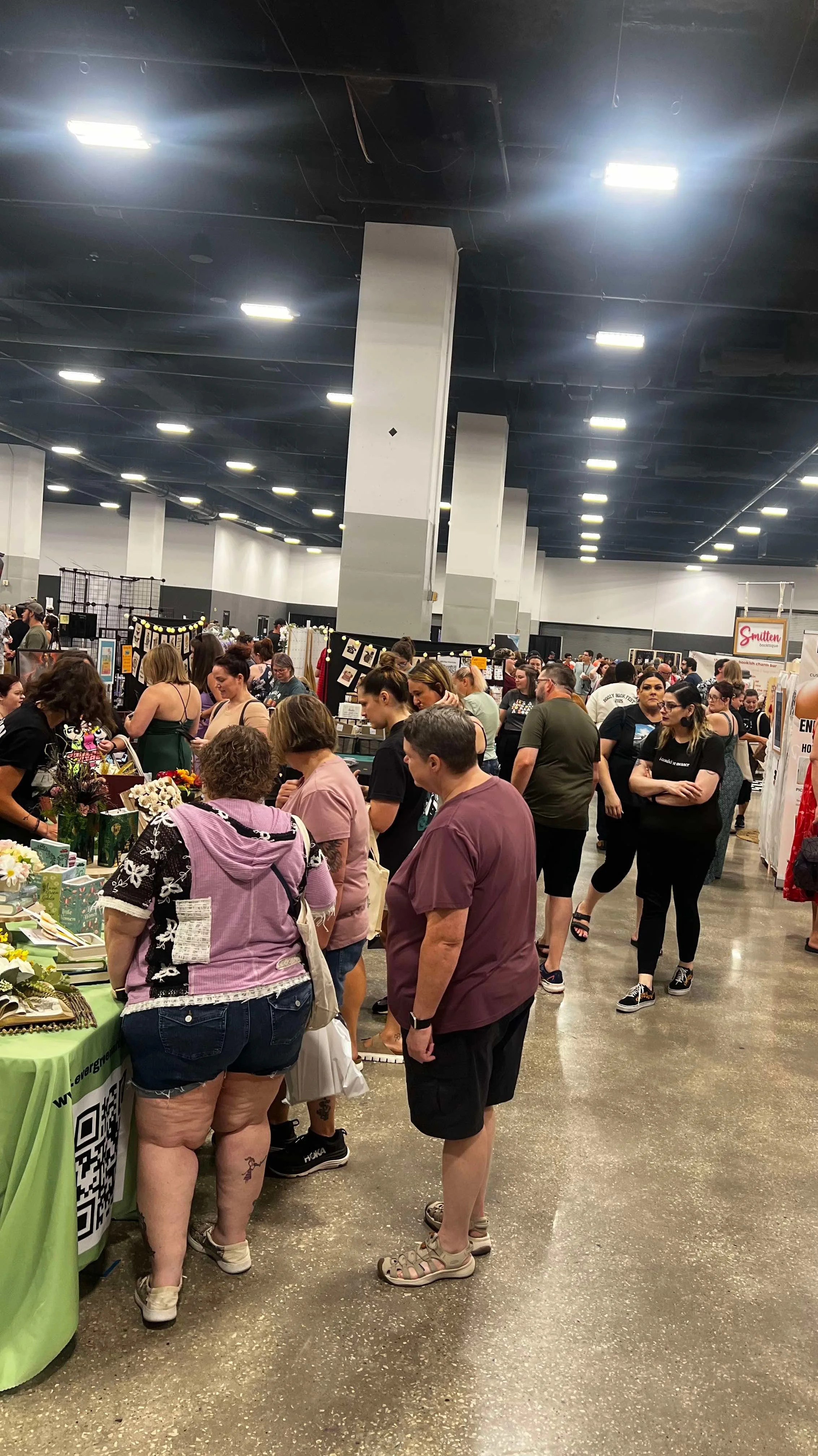 Crowd of people shopping at an indoor market or convention with various booths and tables displaying items.