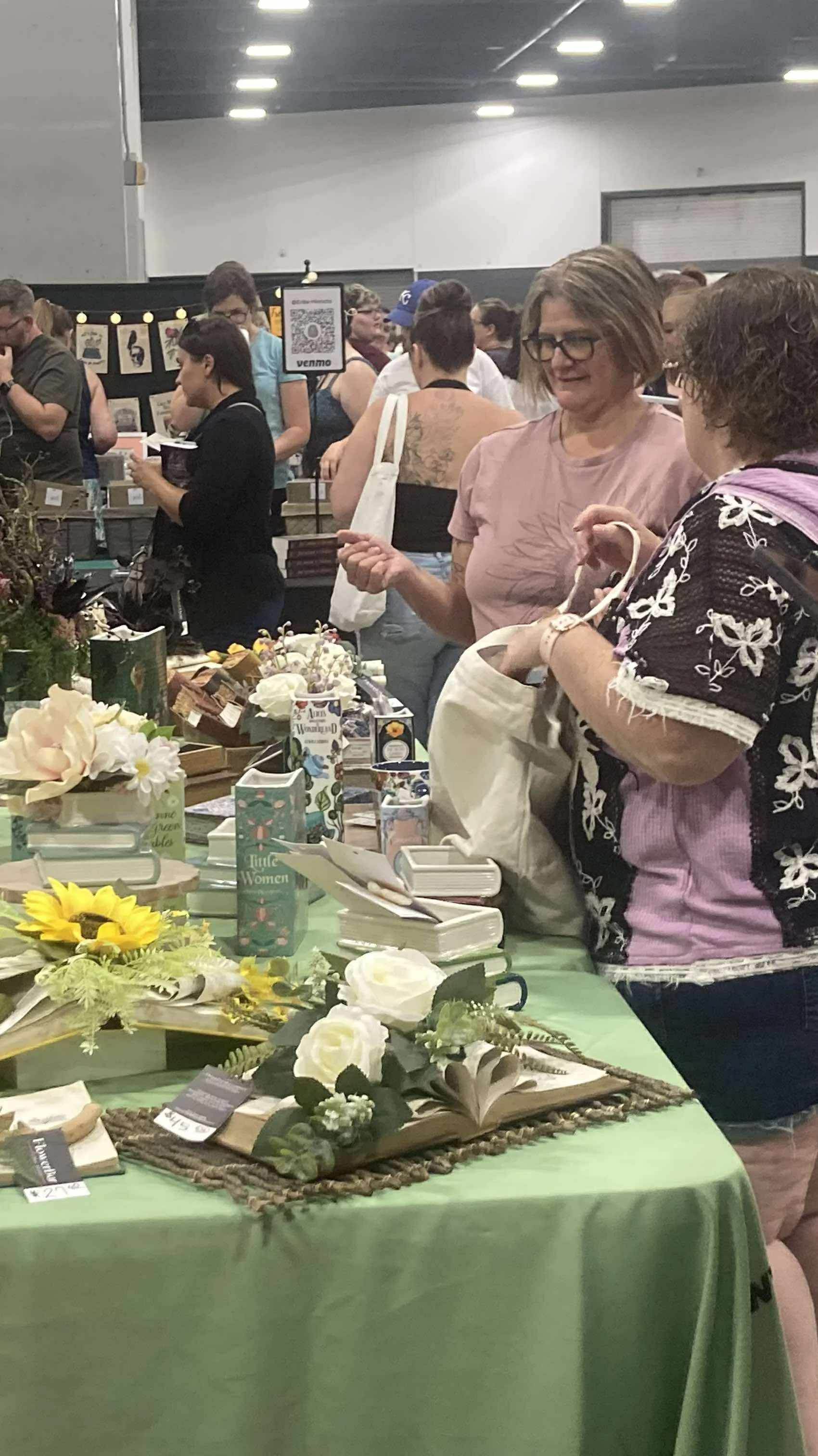 People browsing flower and home decor items at an indoor market or fair, with tables displaying artificial flowers, books, and decorative items.