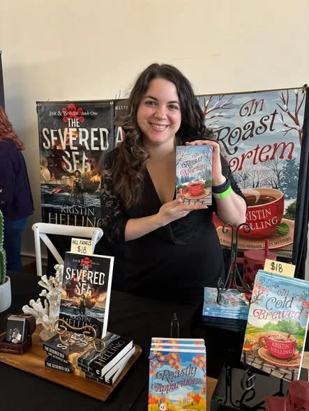 Woman smiling at a book signing event, holding a copy of her book titled "A Cold Brewed Mystery," with books and promotional materials on display.