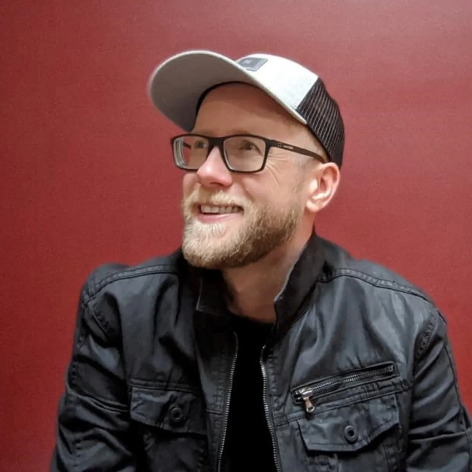 A smiling man with glasses and a beard wearing a black leather jacket and a gray and black cap, sitting against a red background.
