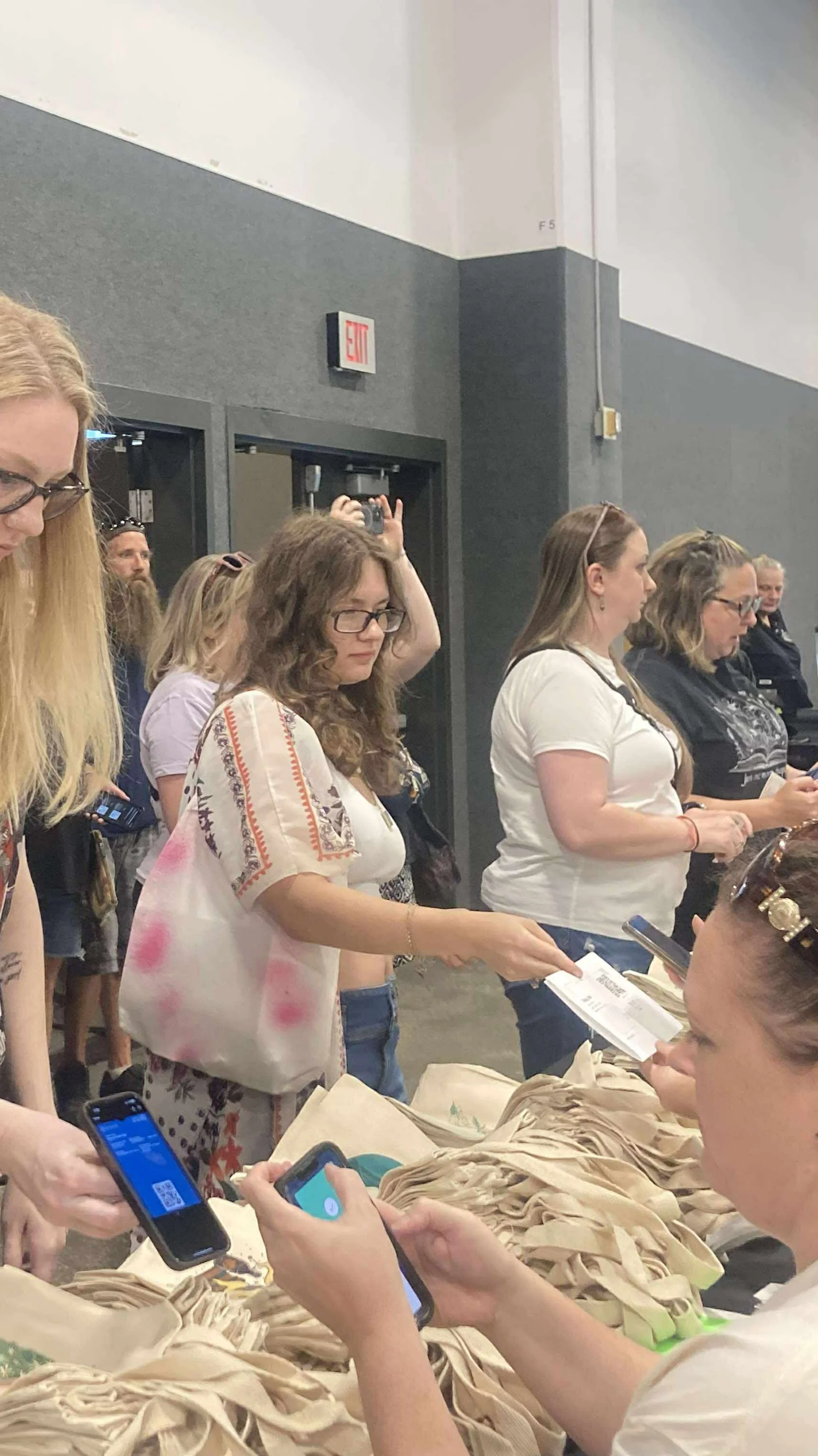Several people standing in a line in an indoor setting, many holding or looking at their phones, with beige fabric or paper items on a table in front of them.