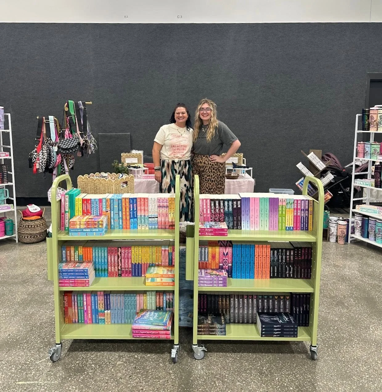 Two women standing behind a table at a book fair, surrounded by bookshelves filled with books, with merchandise like handbags and coffee mugs on display.