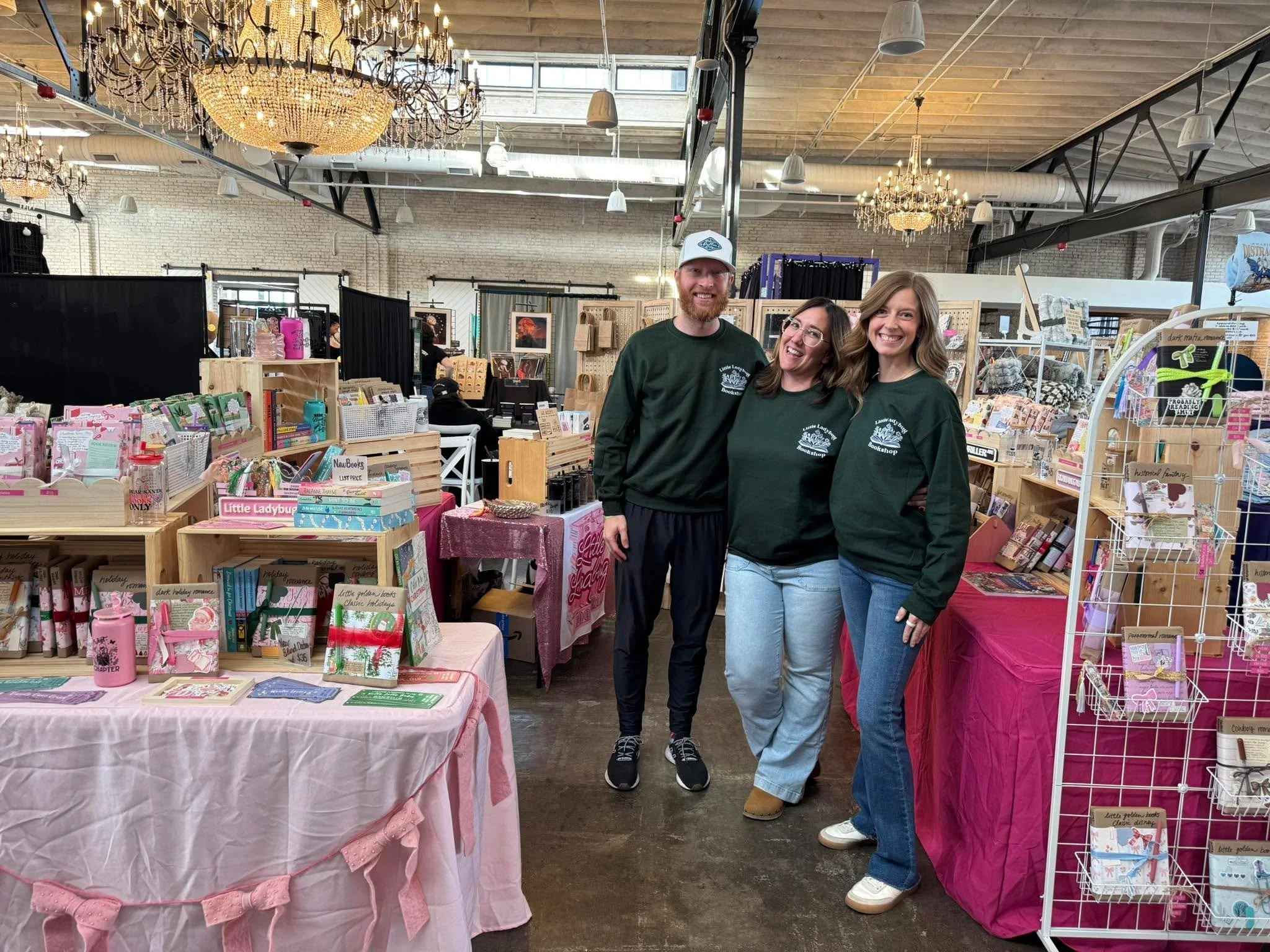 Three people smiling and standing behind a display table at an indoor market or craft fair, with shelves of books and various merchandise visible around them.