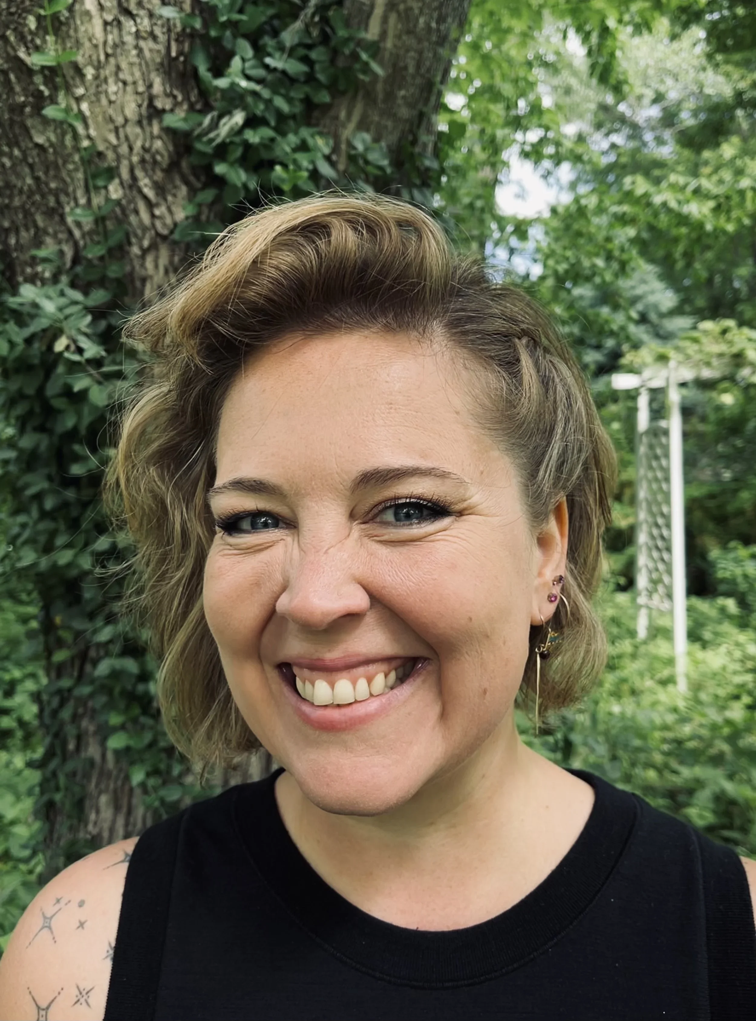 Close-up of a smiling woman with short, wavy blonde hair outdoors in front of a tree and green foliage, wearing a black sleeveless top and earrings.