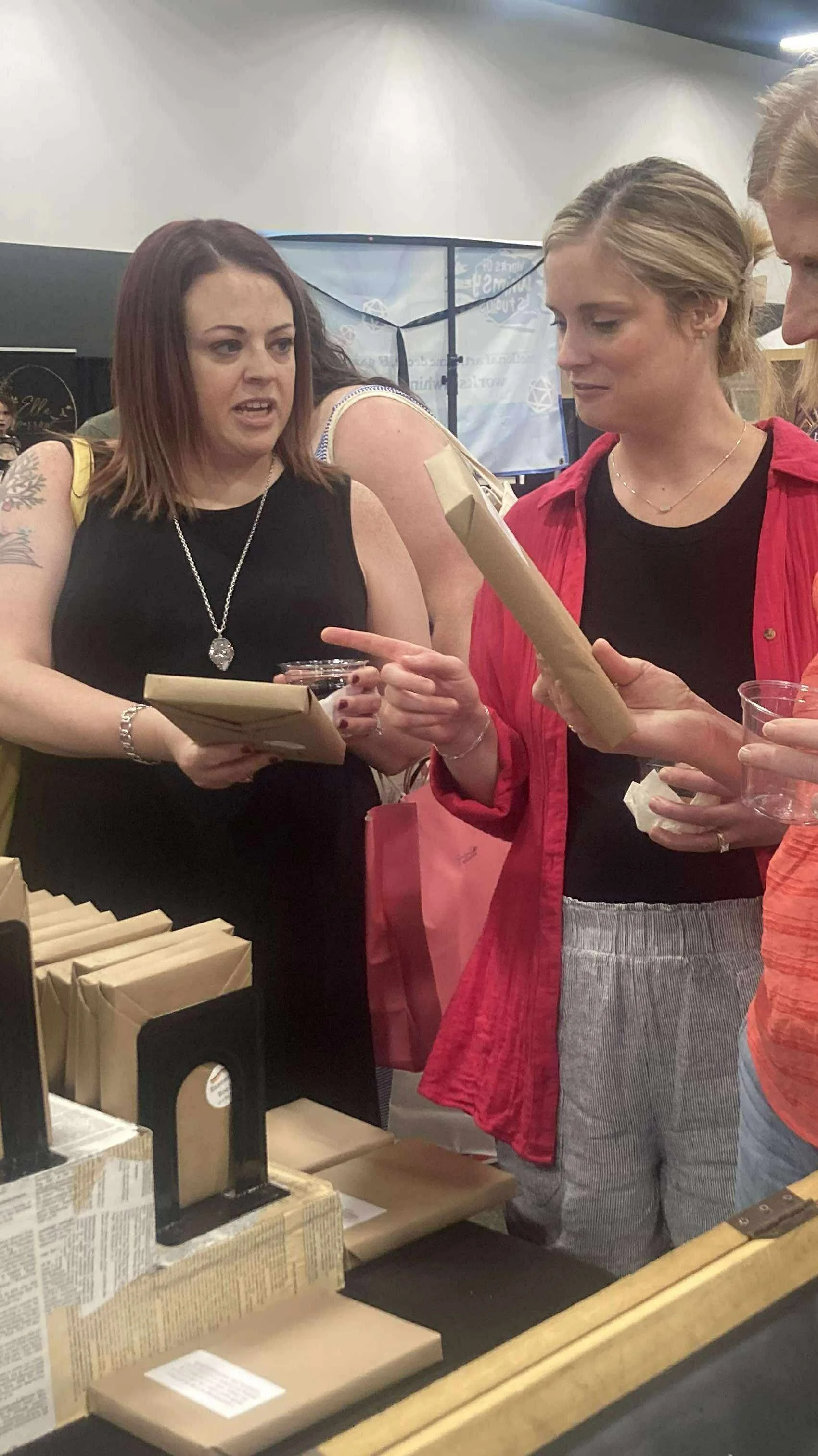 Two women are at a table, looking at items in their hands, with retail packaging on the table, in a store or event setting.