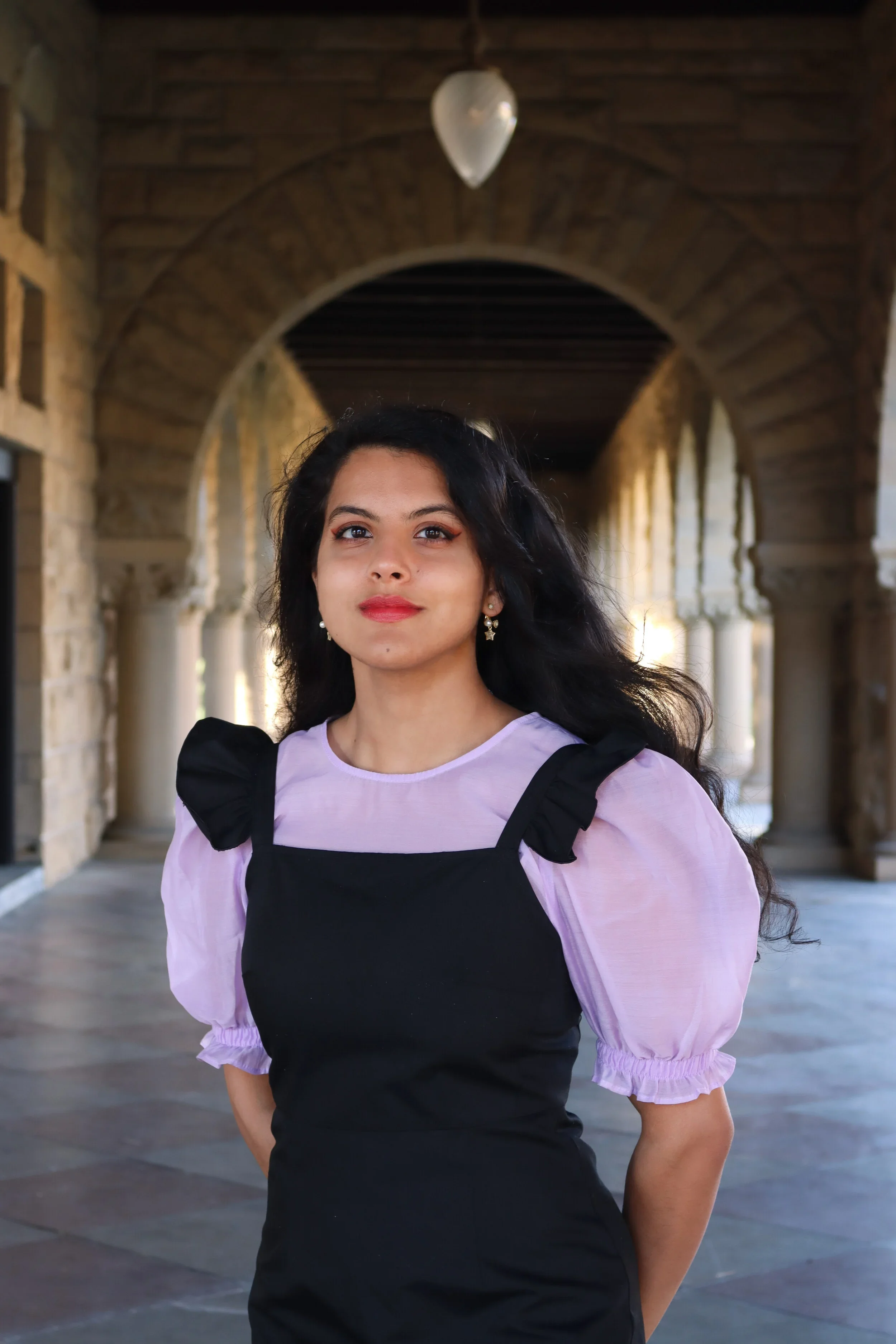 A young woman with long dark hair and earrings, wearing a lavender blouse with puffed sleeves and a black pinafore dress, standing under an arched stone walkway with columns.