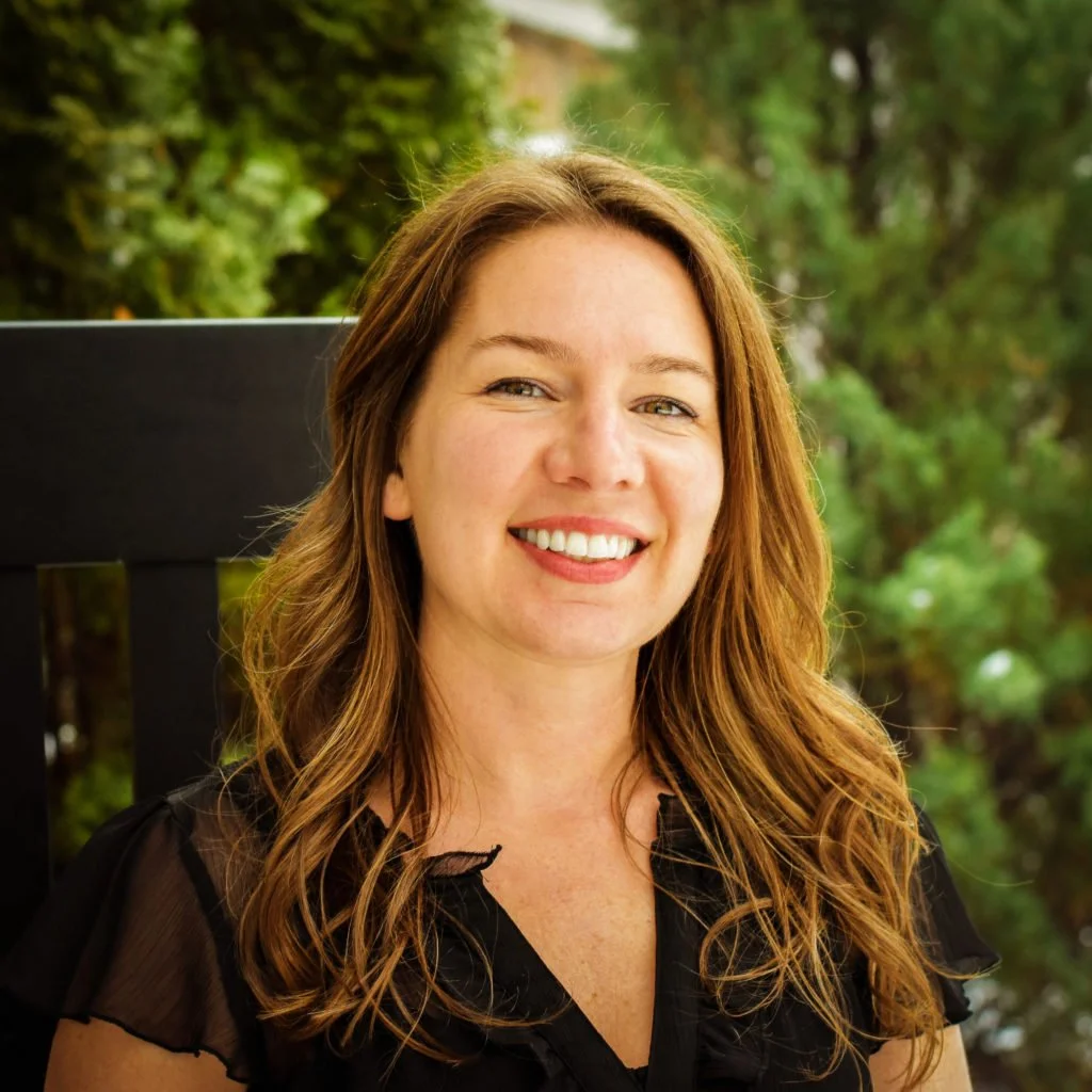 A woman with long, wavy, auburn hair smiling outdoors with trees in the background.