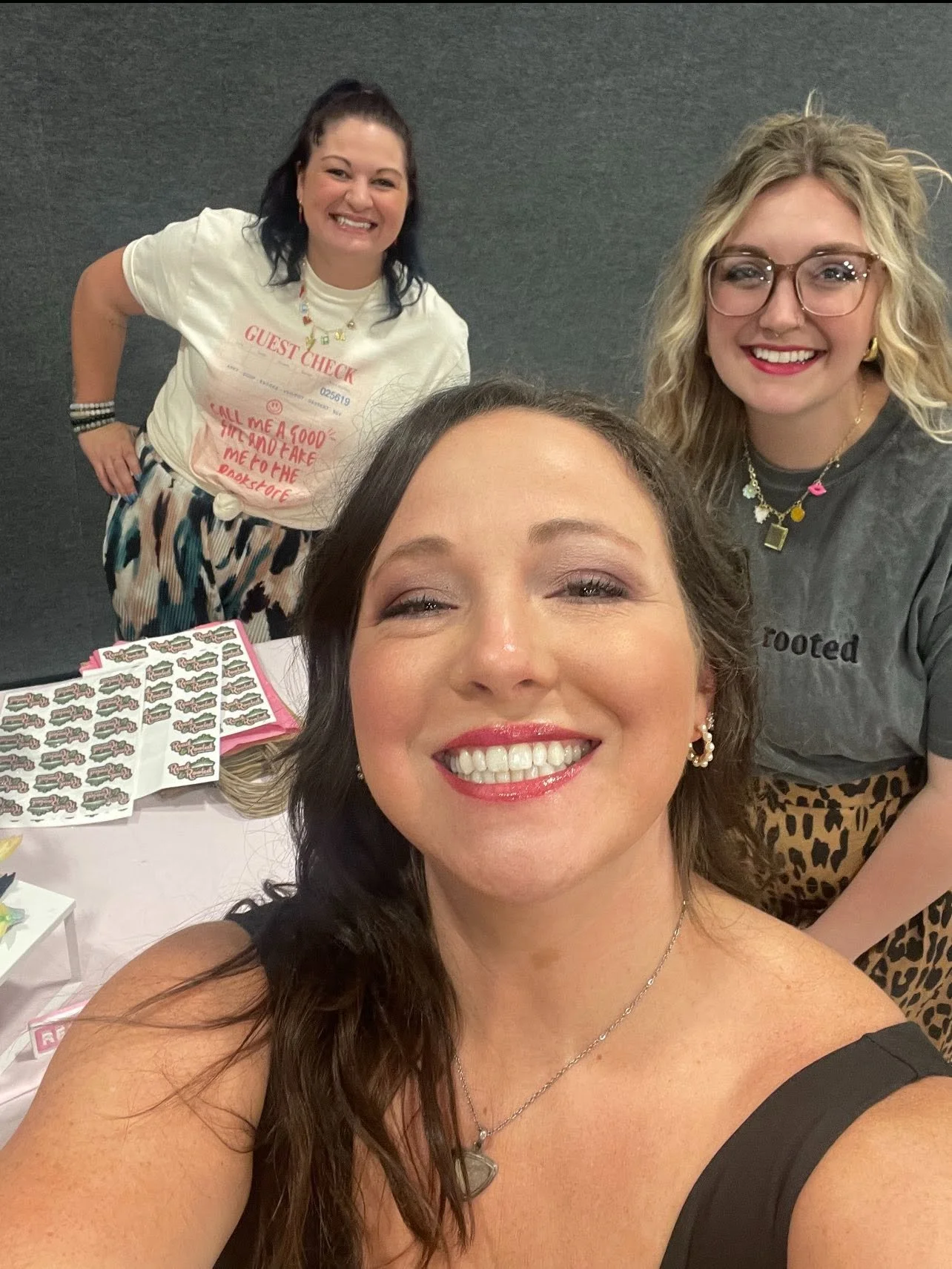 Four women smiling in a group, one woman taking a selfie with the others in the background. There are stickers, paper, and promotional items on the table.
