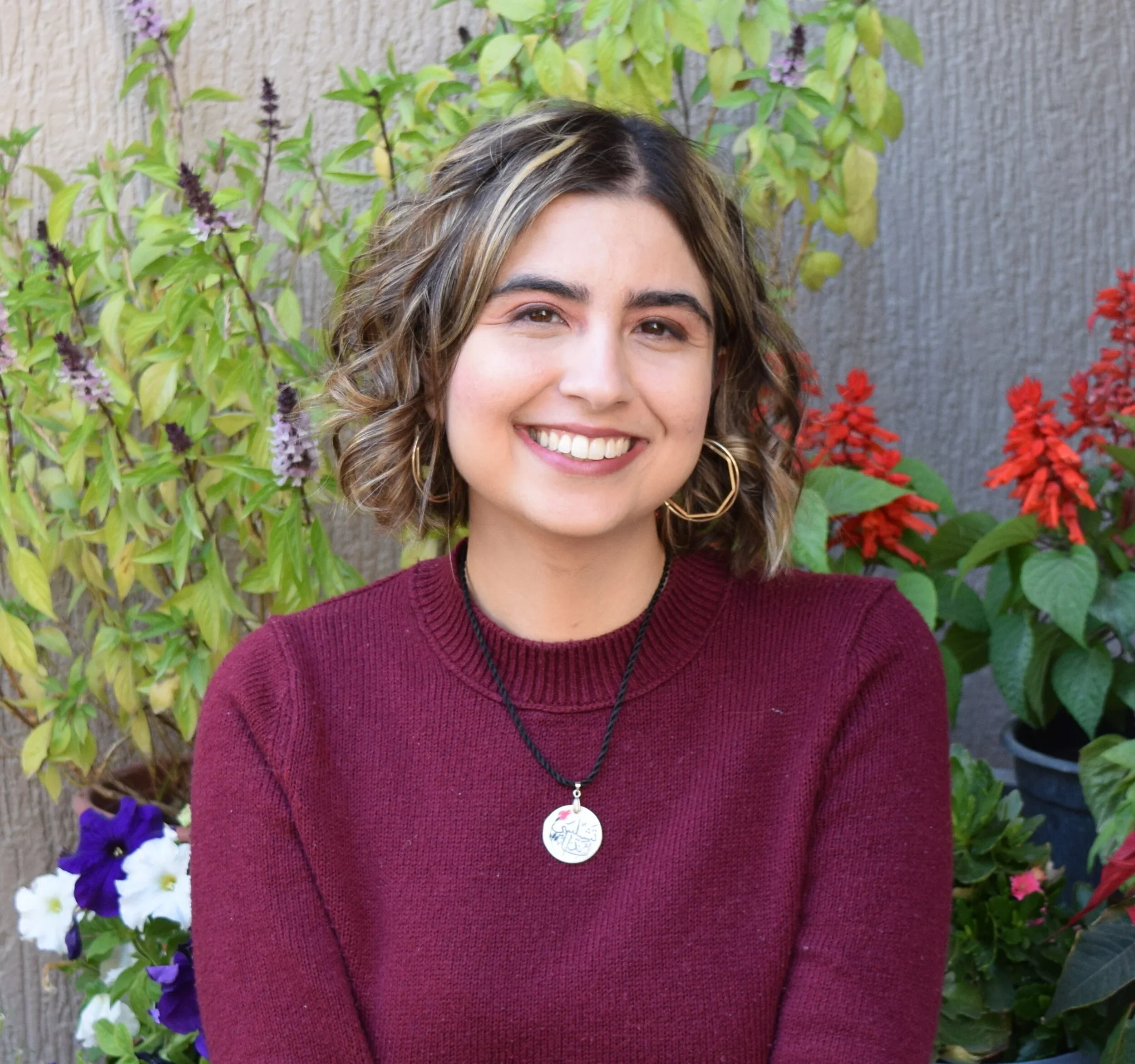 A woman with curly hair and hoop earrings smiling in front of a garden with various flowering and green plants.