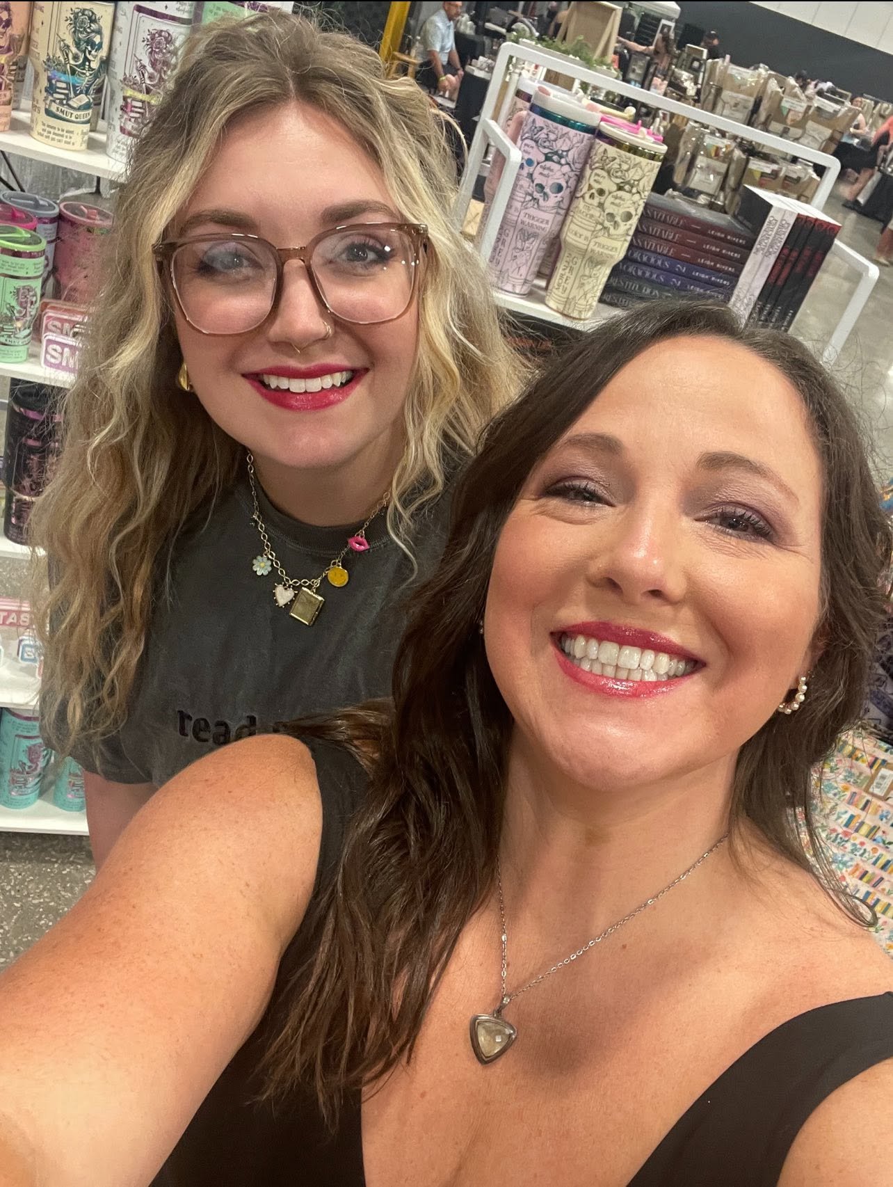 Two smiling women taking a selfie at a book or comic convention, with shelves of books and colorful merchandise in the background.