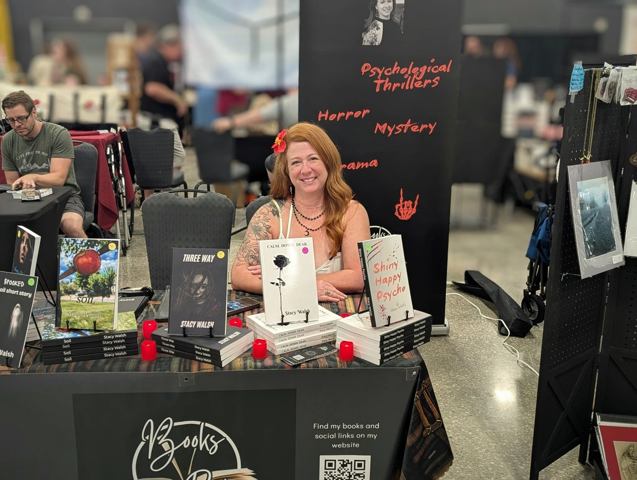 A woman with red hair and tattoos smiling at a book signing event, seated at a table with books displayed in front of her. Behind her, a black sign reads 'Psychological Thrillers, Horror, Mystery, Drama,' with a small photo of her. The background sho