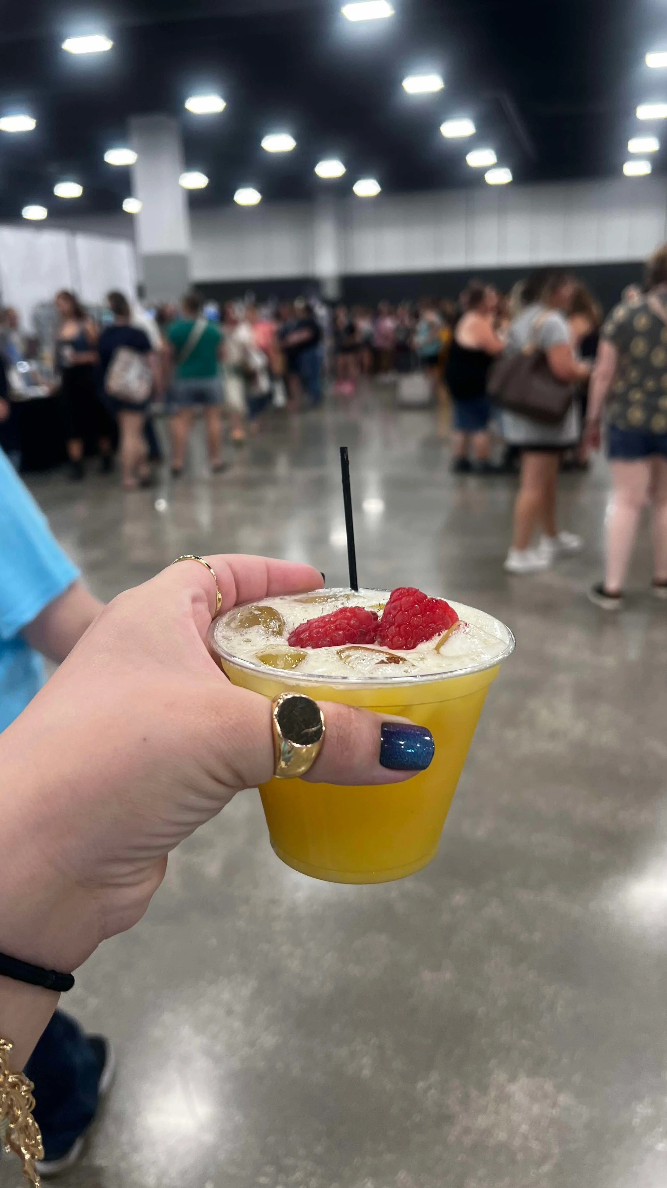 A person holding a yellow fruit smoothie topped with raspberries and green grapes at an indoor event with a large crowd in the background.