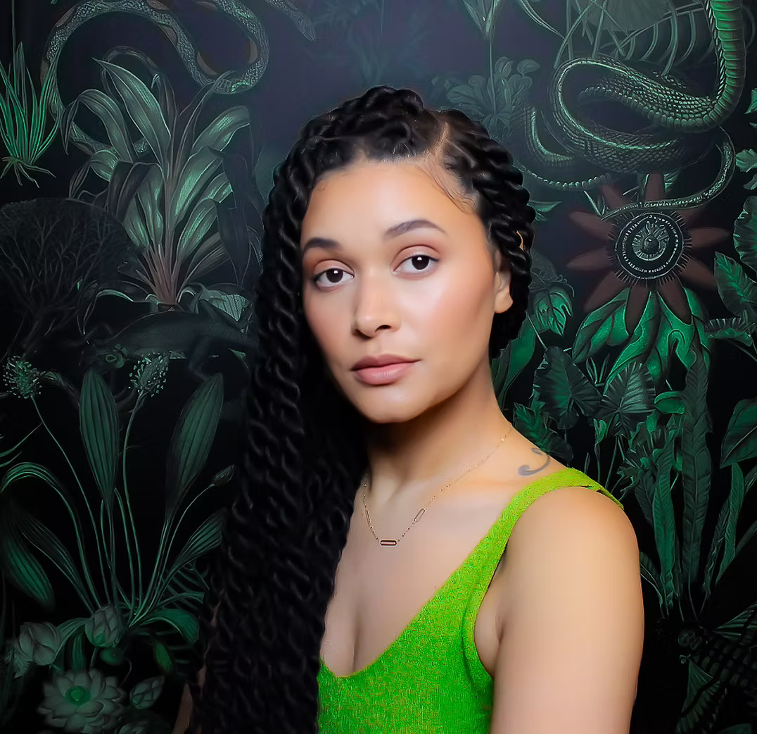 A young woman with long, black, twisted braids wearing a bright green sleeveless top, standing in front of a dark jungle-themed background with green plants and snakes.