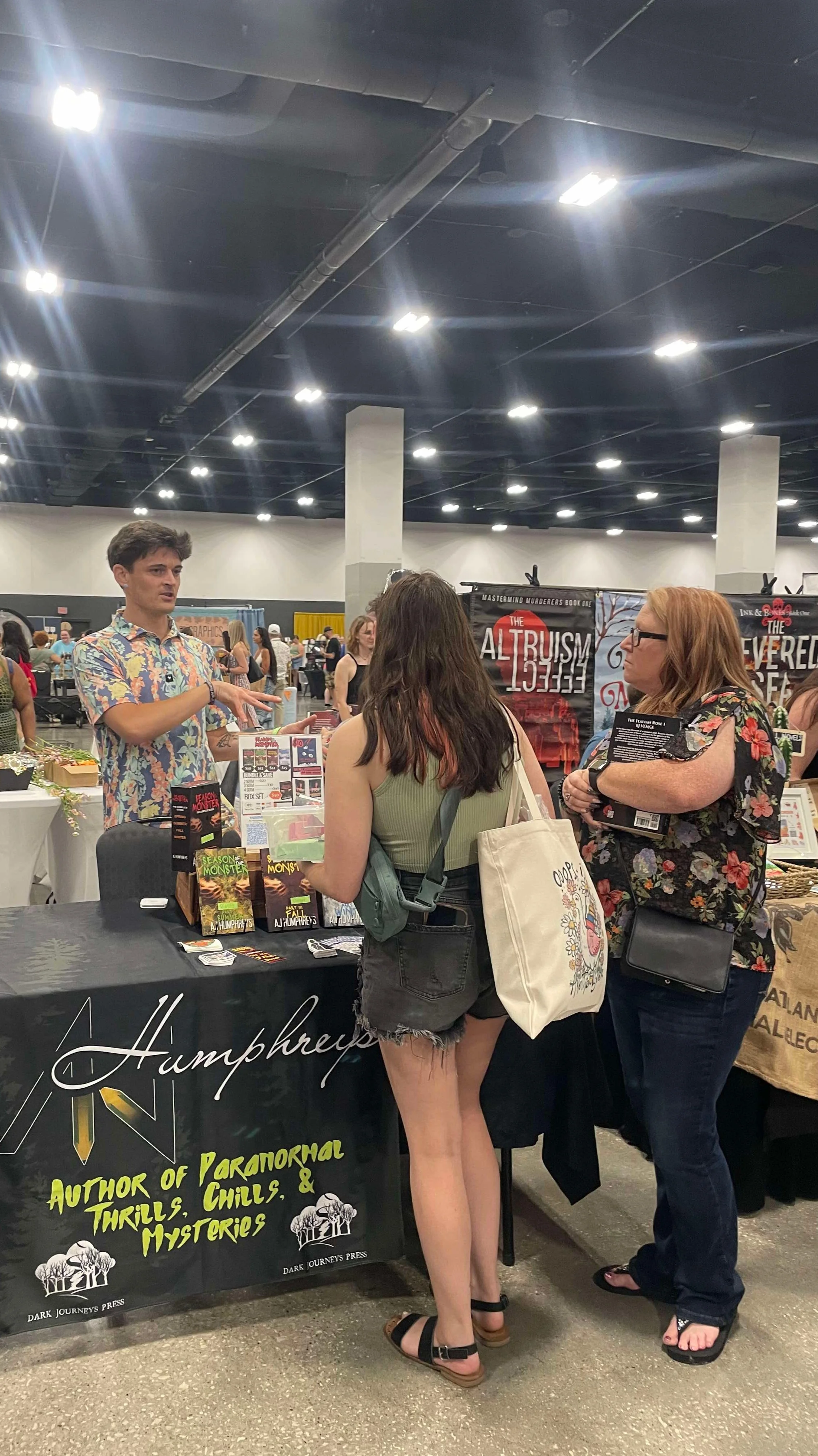 A man in a colorful, floral shirt is talking to two women at a booth at a convention or expo. The booth has books and merchandise related to horror, paranormal thrills, and mysteries, with a black tablecloth and banners displaying the author's name.