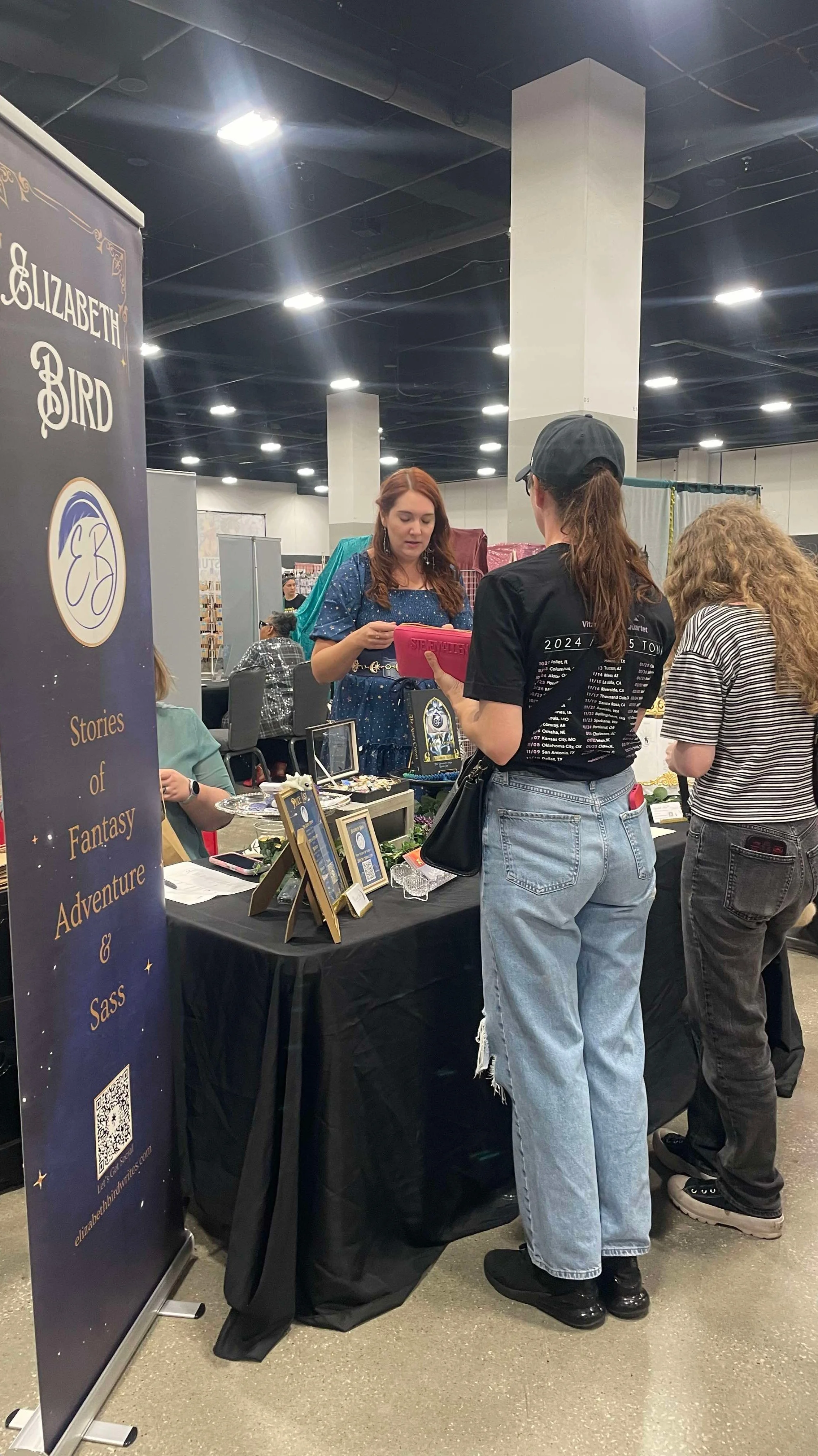 Two women are talking at a booth with jewelry and Fantasy-themed merchandise, with a large Elizabeth Bird banner on the side. The scene takes place in an indoor convention or fair with multiple booths and people in the background.