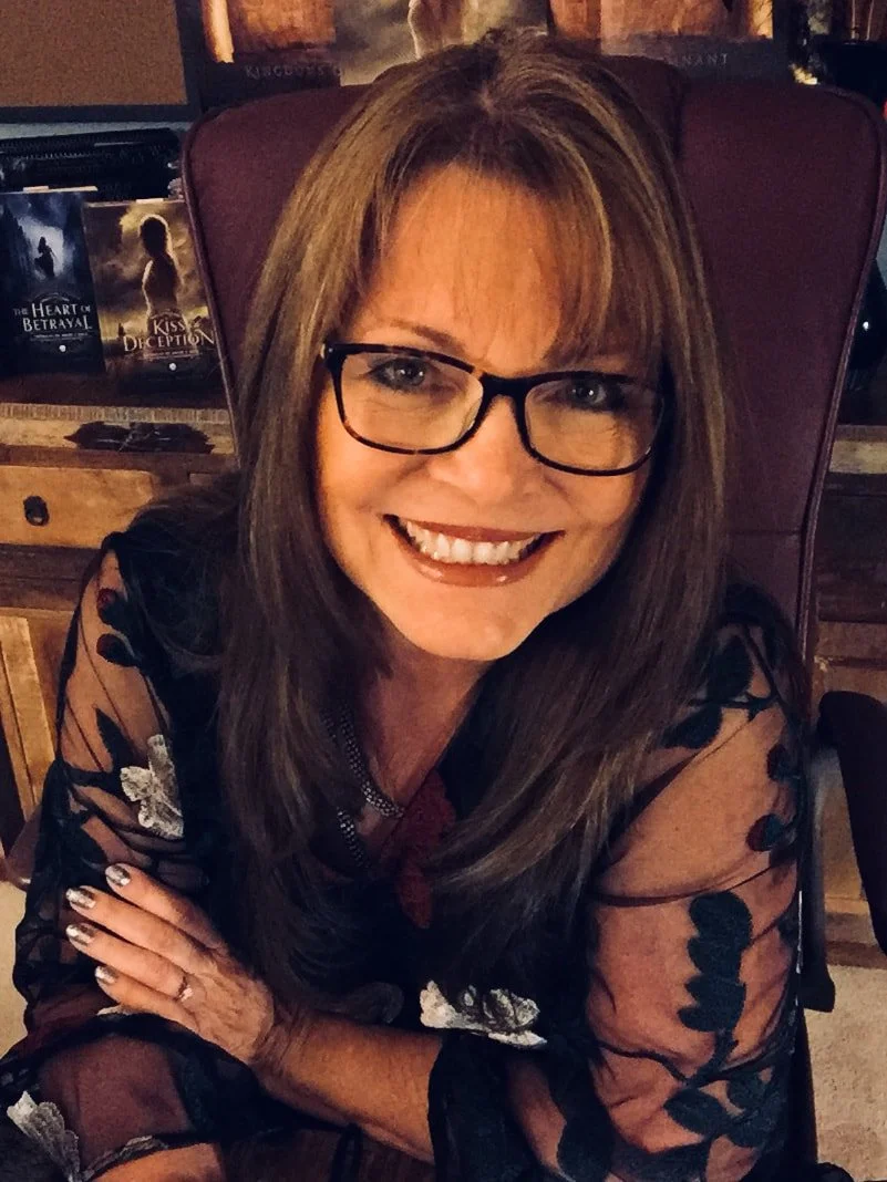 A woman with shoulder-length brown hair, glasses, and a friendly smile, sitting at a wooden table in a cozy, dimly lit room; she is wearing a black floral-patterned sheer top and has her arms crossed on the table.