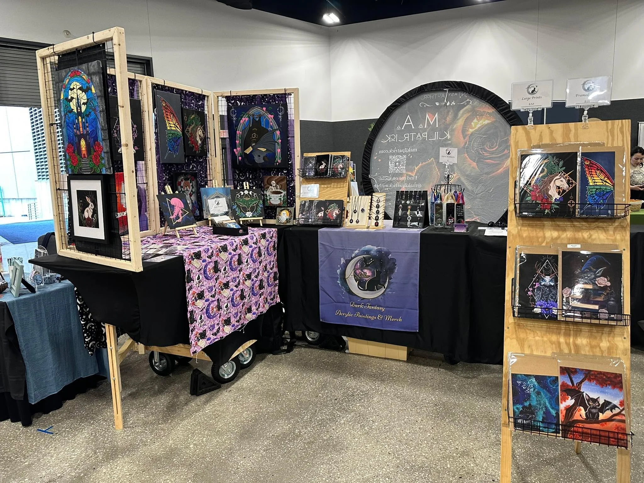 An art booth at an indoor event displaying colorful acrylic paintings of unicorns, dragons, and fantasy scenes on black backgrounds. Artwork is on wooden and wire frames, with some small framed pieces on tables.