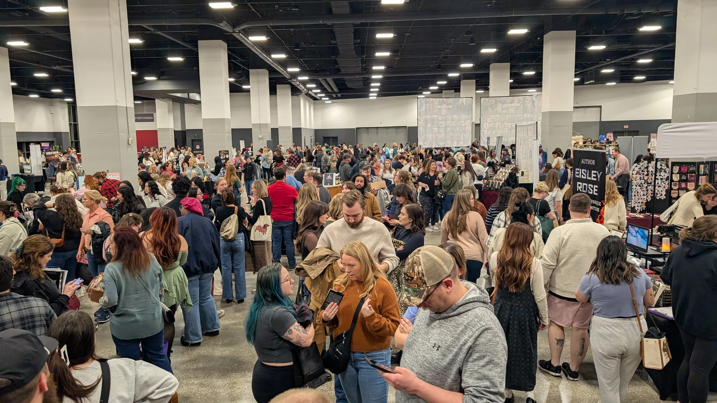 Crowded indoor event with many people browsing vendor tables and displays, some engaged in conversations and others looking at their phones.