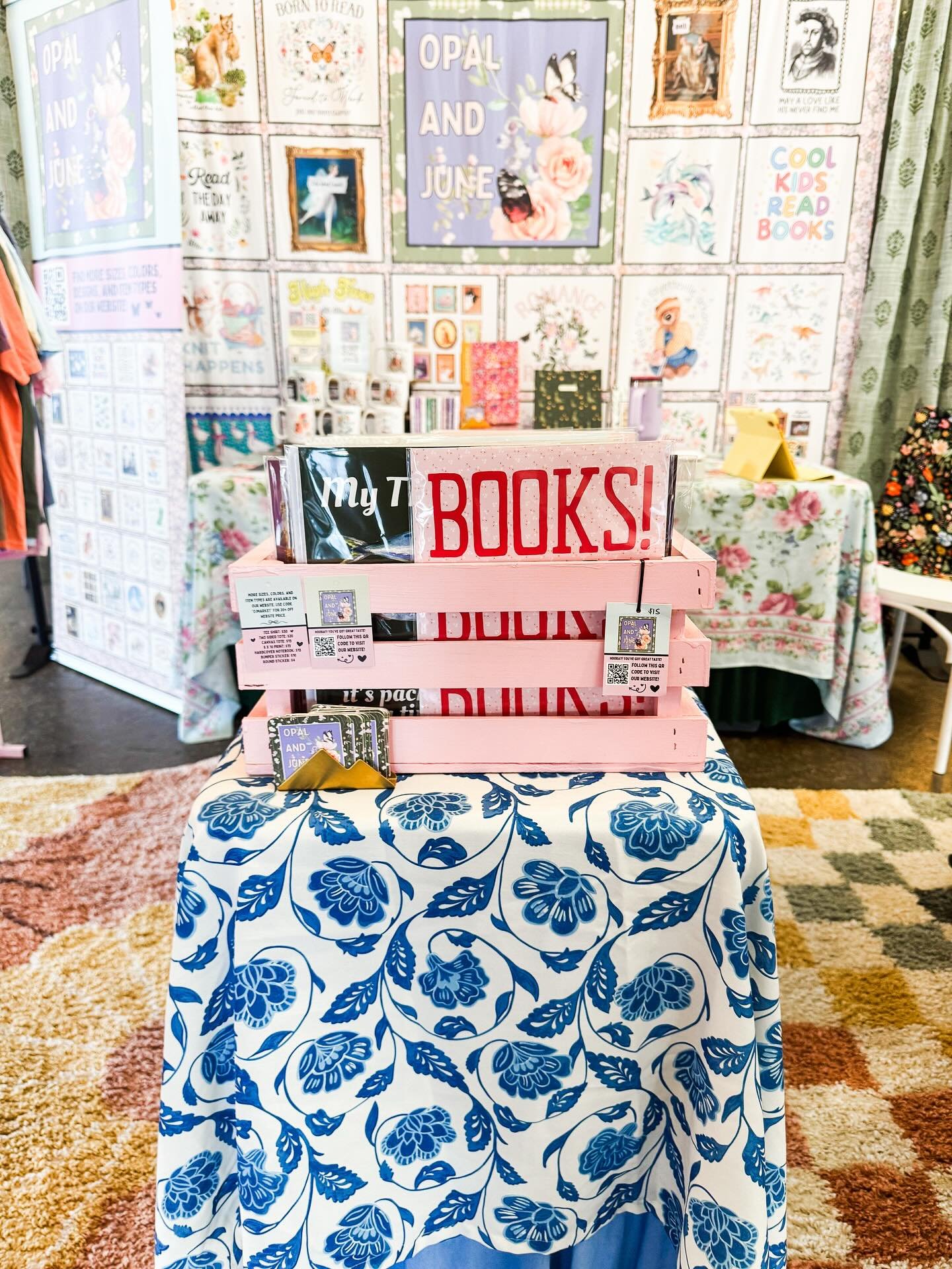 A display table covered with a blue and white floral tablecloth, holding a pink wooden crate with signs that say 'My T Books!' and 'Books!'. Behind the table, there is a wall with colorful artwork and posters, including one that says 'Opal and June' 