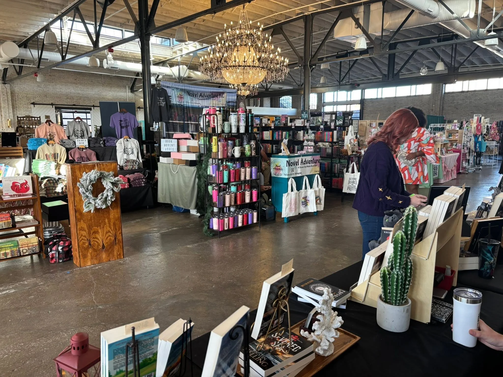 Inside a bookstore or market with shelves of books, clothing, and accessories. A large chandelier hangs from the ceiling. Customers browse the items, including a woman with red hair and a blouse with stars.