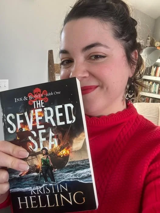 A woman with dark hair tied back, wearing a red sweater, holding a copy of the book 'The Severed Sea' by Kristin Helling, inside a room with bookshelves in the background.