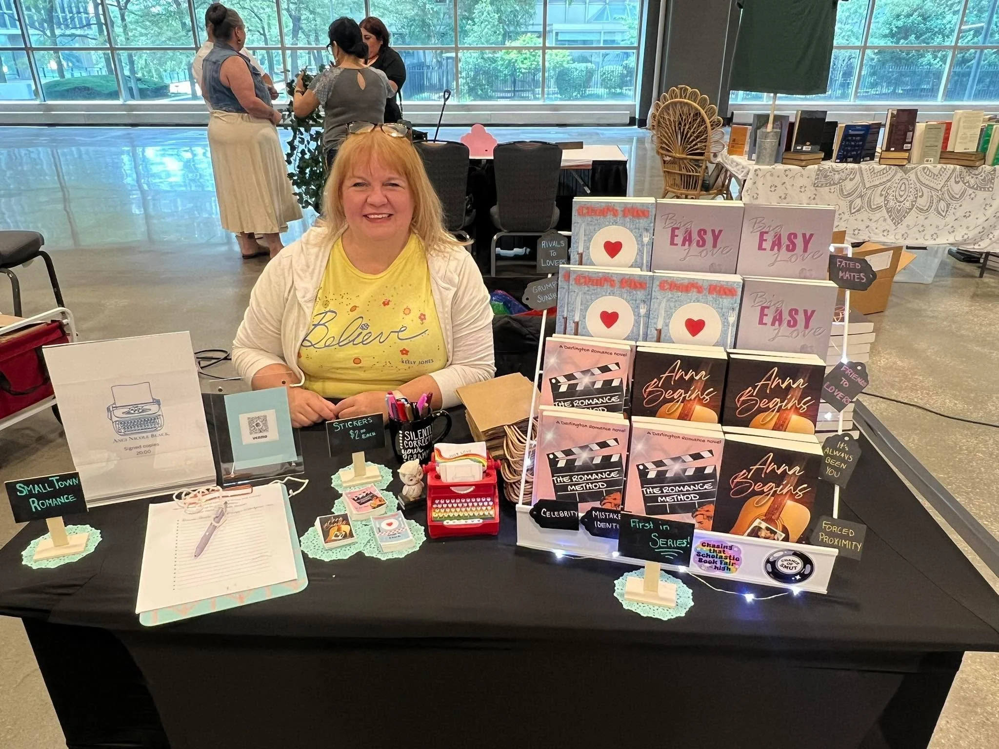Woman sitting at a book table selling romance novels at a book fair, with books, signs, and decorative items on display. Three women are in the background near large windows.
