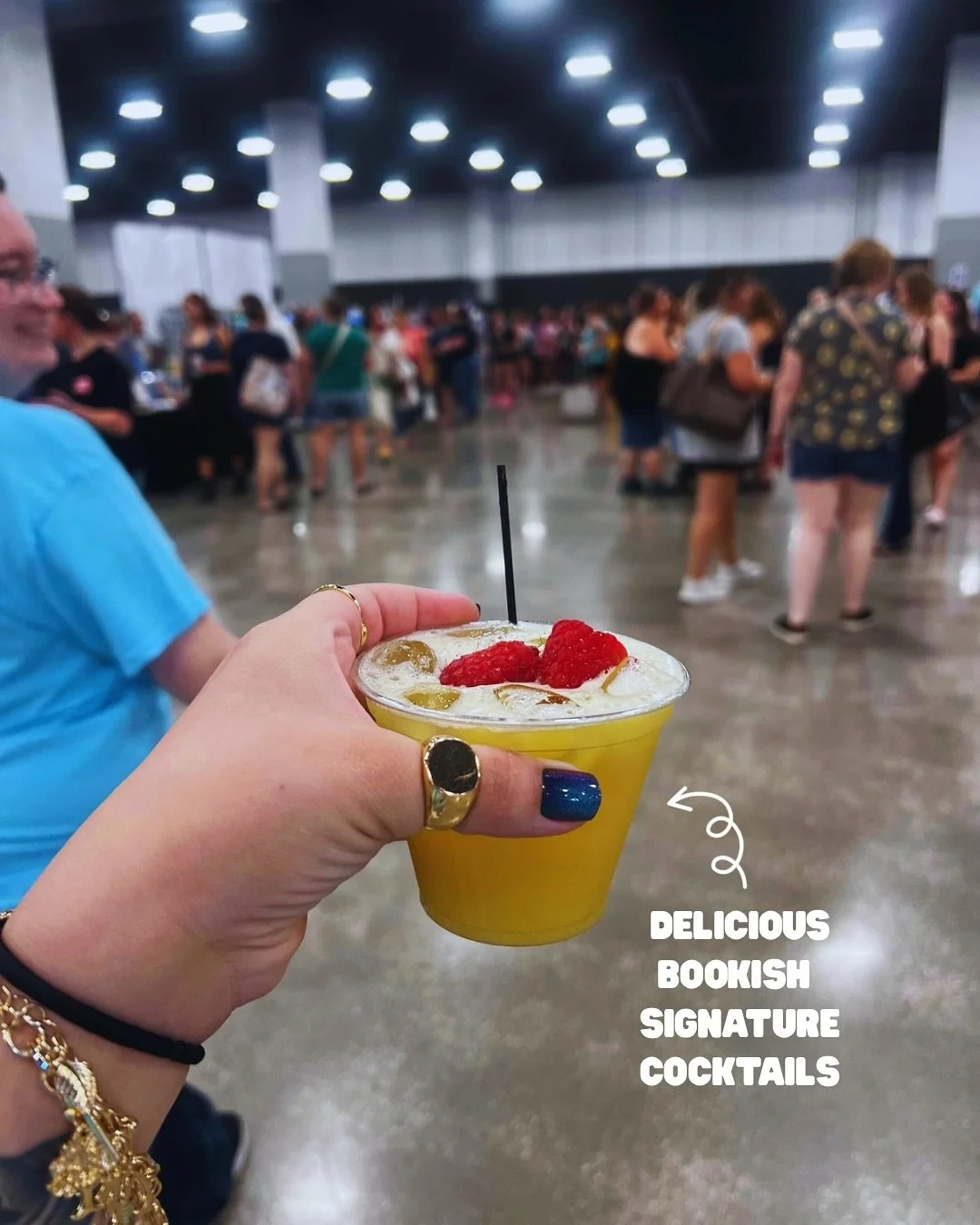 A hand holding a yellow cocktail garnished with raspberries and other fruit in a crowded indoor event space. Text on the image reads 'Delicious bookish signature cocktails.'