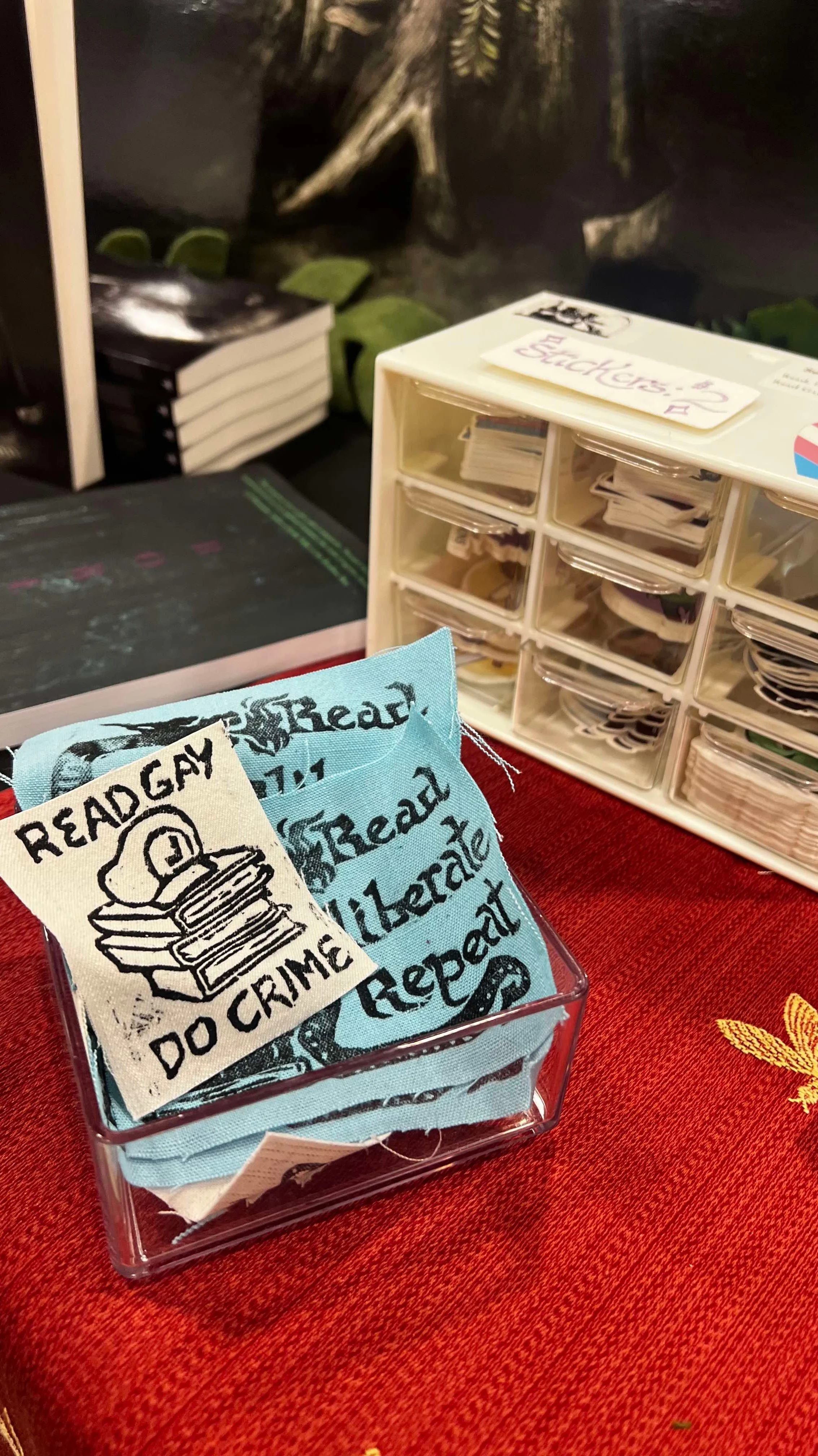 Close-up of a transparent container holding blue fabric patches with crime-themed text and illustrations, placed on a red textured surface.