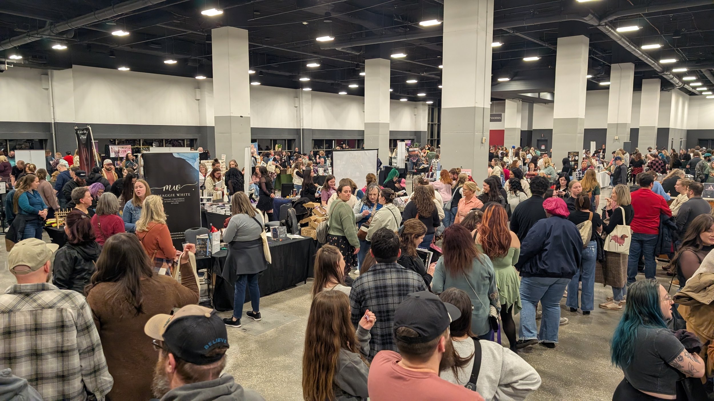 A crowded indoor event or convention with many people browsing booths and tables, some holding shopping bags, in a large open space with high ceilings and bright lights.