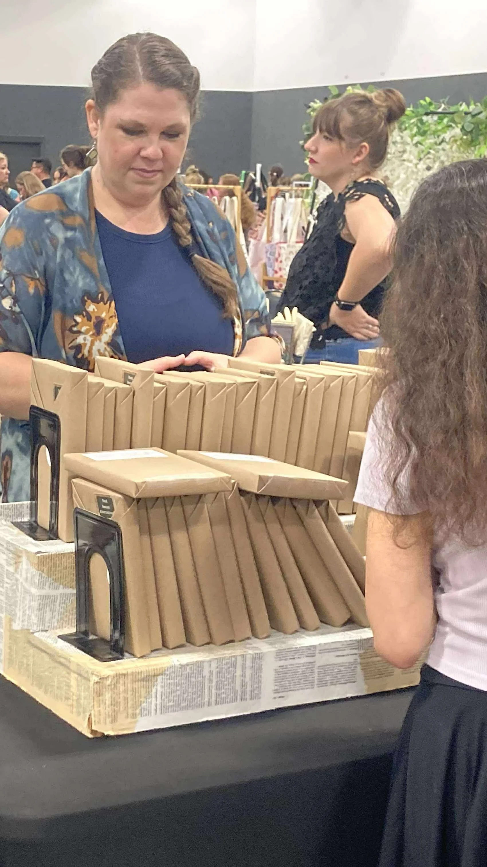 A woman with braided hair wearing a blue top and patterned jacket is at a market or event, looking at items on a table with brown paper packages. There are people in the background, some browsing or standing, and the setting looks like an indoor even